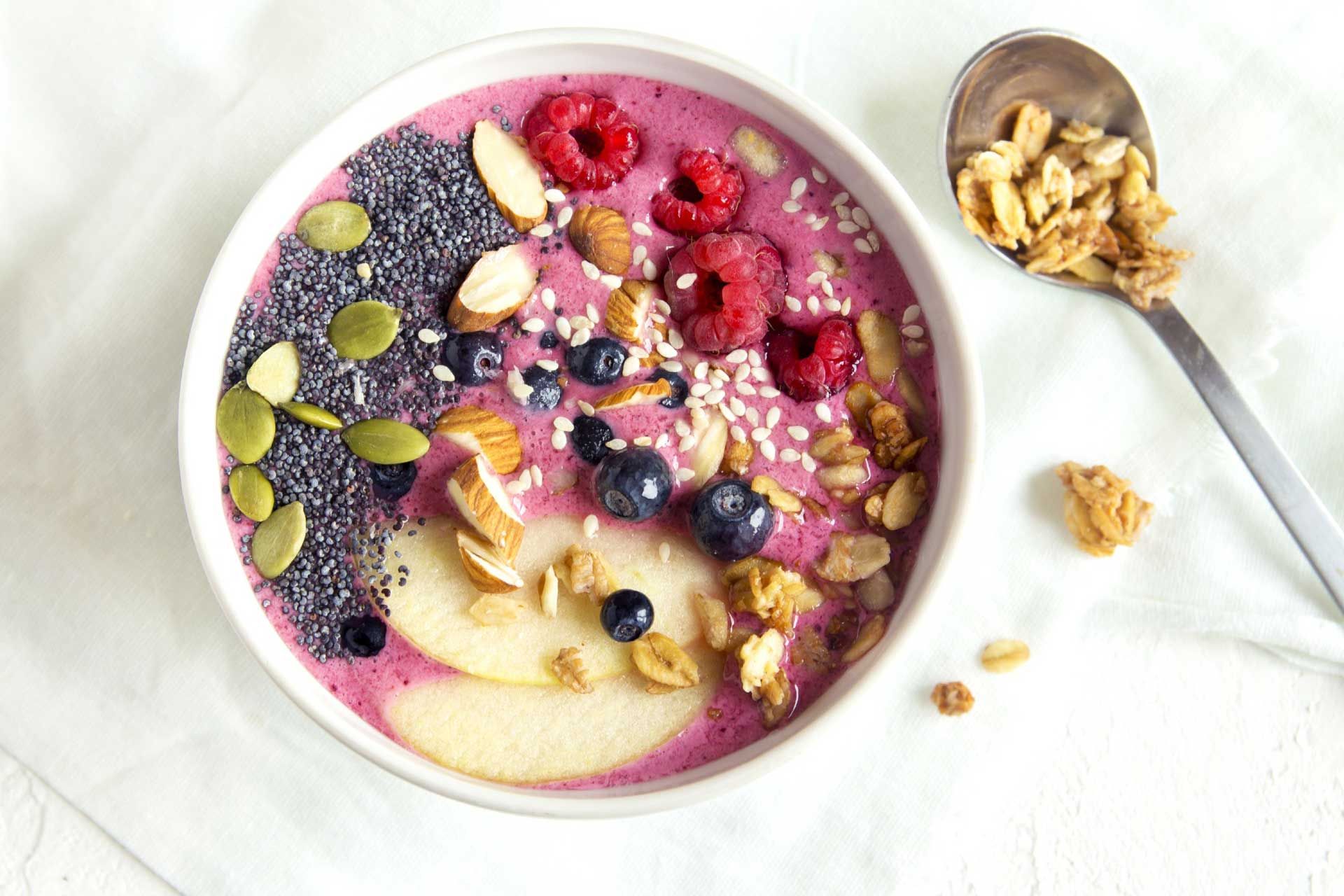 Smoothie bowl with fresh berries, chia seeds, nuts, and granola, next to a silver spoon.