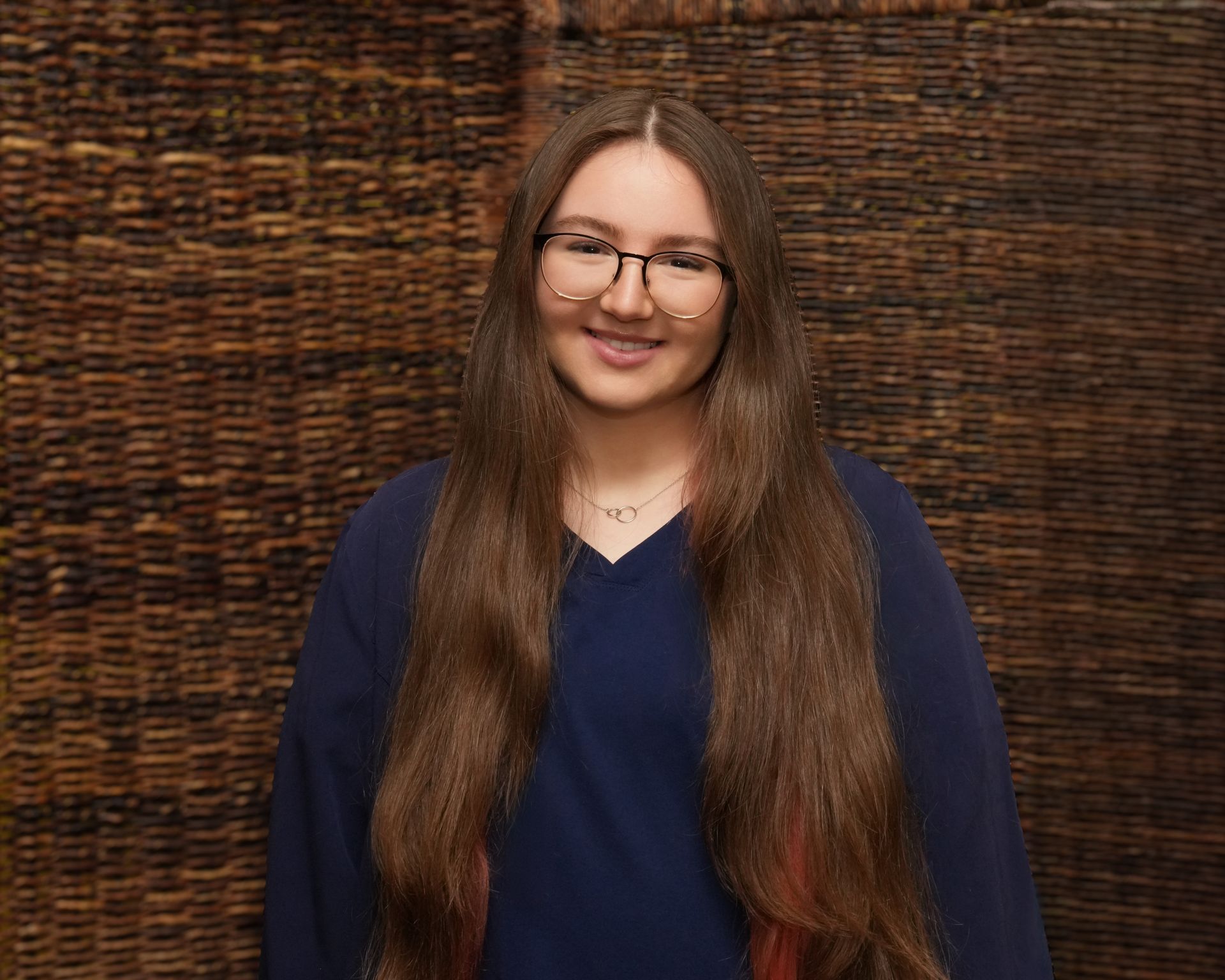 Smiling person with long brown hair and glasses, wearing a dark blue top against a woven brown backdrop
