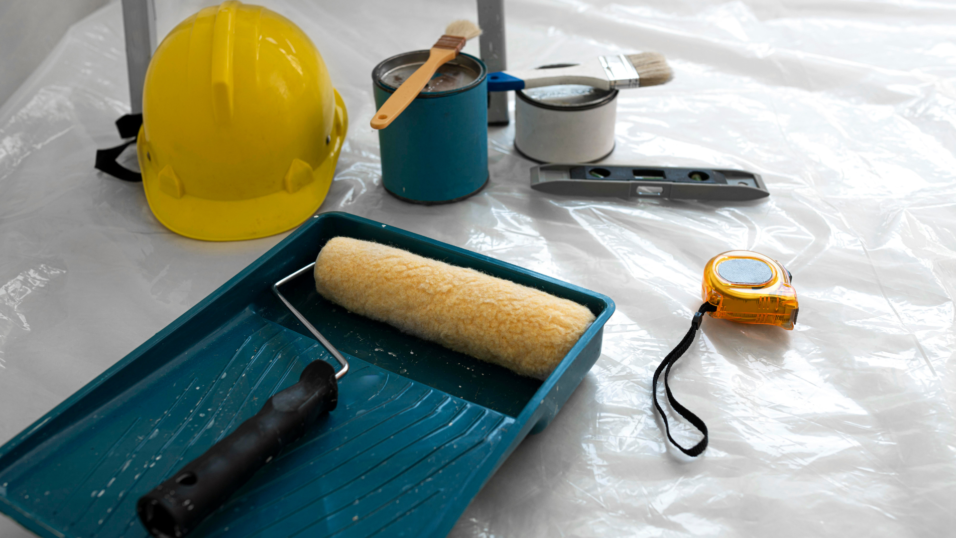 A yellow hard hat , a paint roller , a measuring tape , and paint cans are on a plastic sheet.