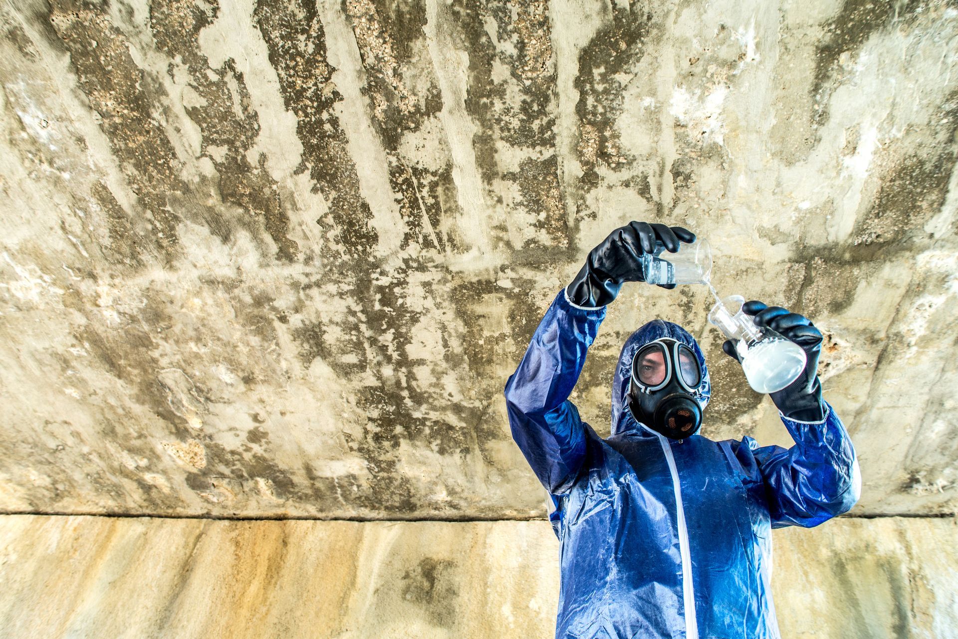 A man in a gas mask and gloves holds a bottle, prepared for emergency mold remediation tasks.