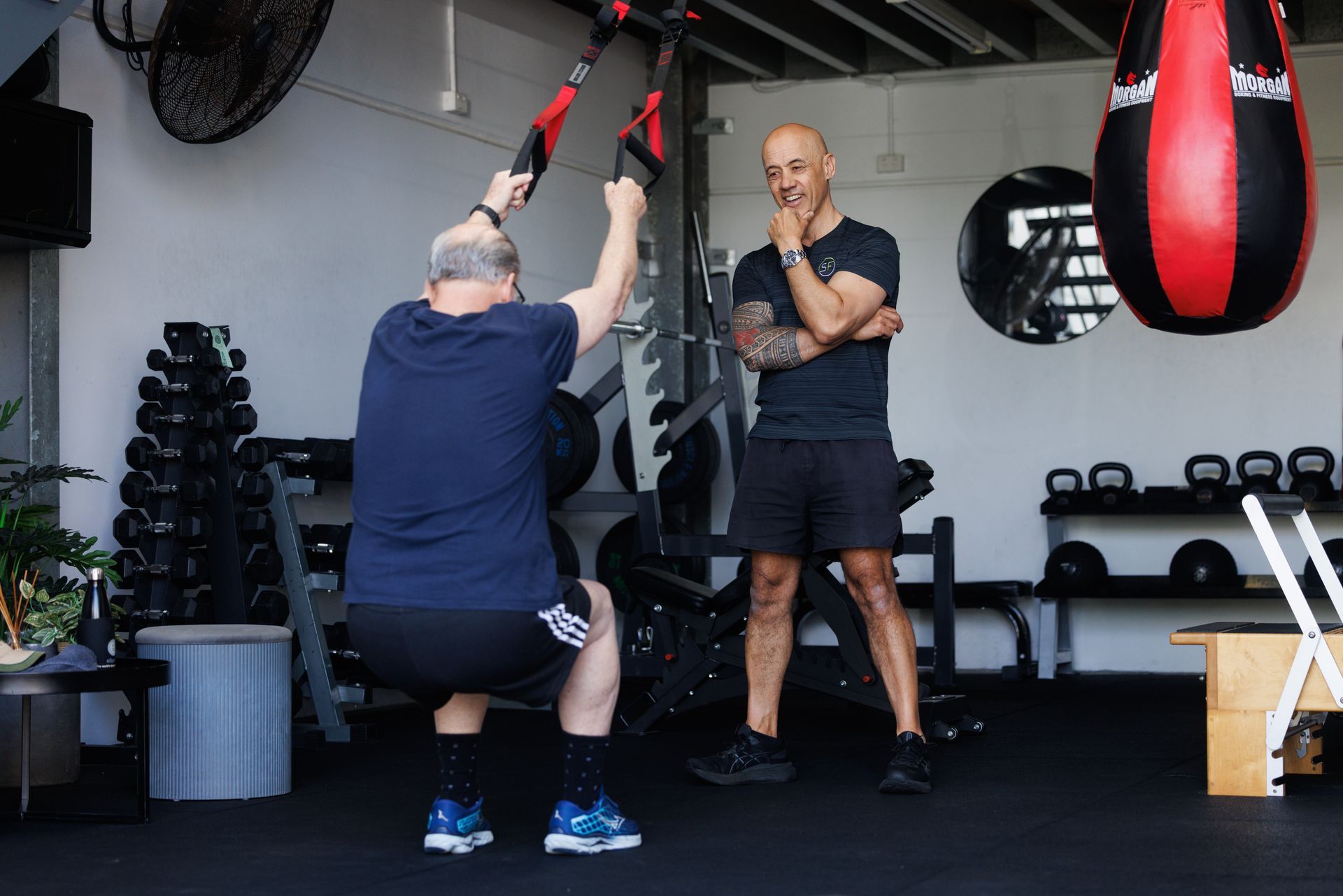 Man doing TRX squats with trainer observing in a gym. Black floor, weights, punching bag.