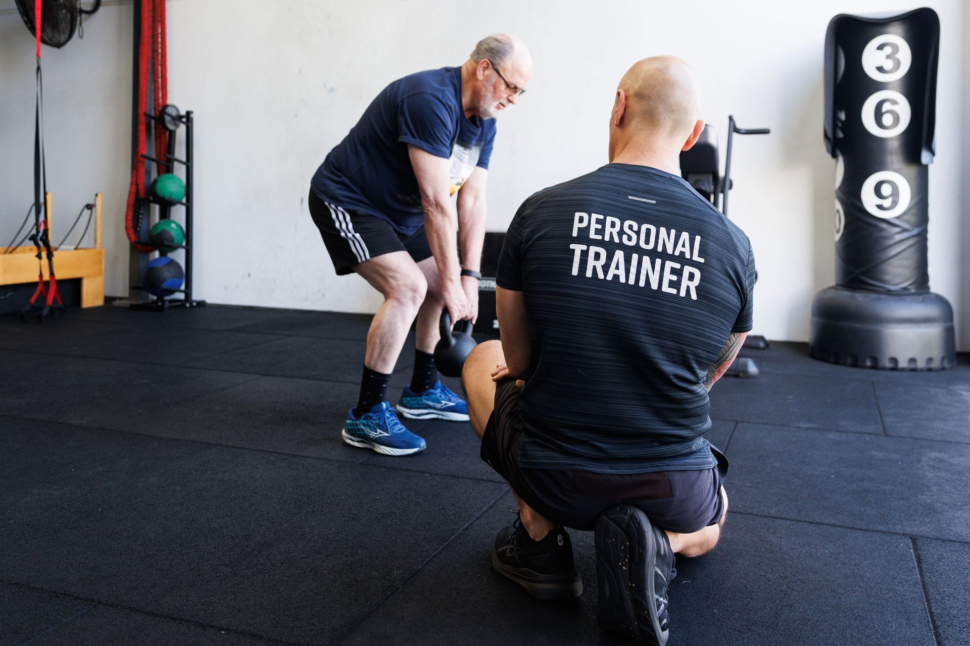 Man lifting kettlebell watched by personal trainer in a gym.