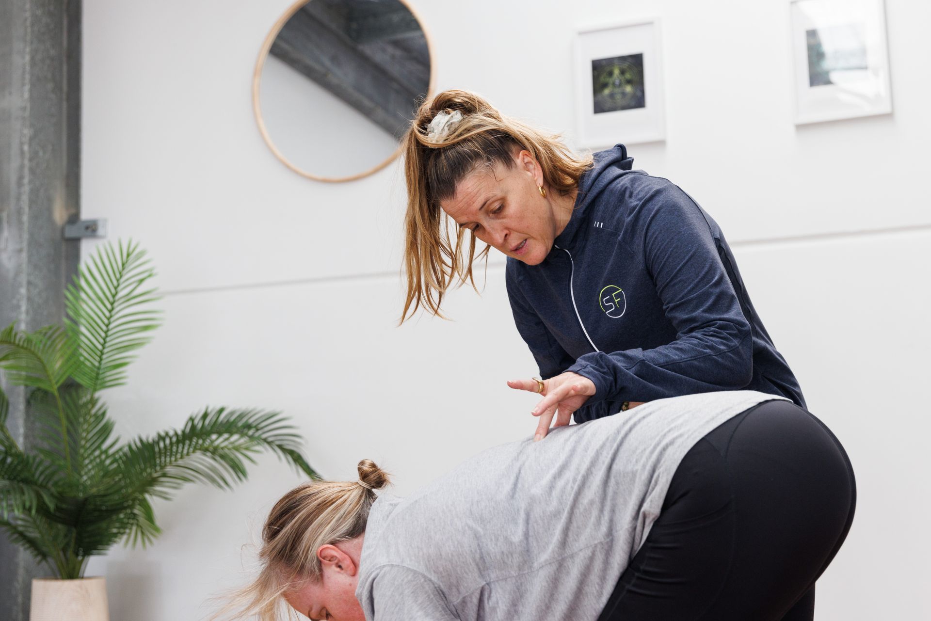 Woman in hoodie assisting another woman's yoga posture in a studio with plants and a mirror.