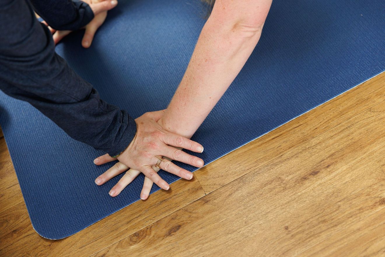 Hands clasped together on a blue yoga mat, one arm extended. Wooden floor in the foreground.