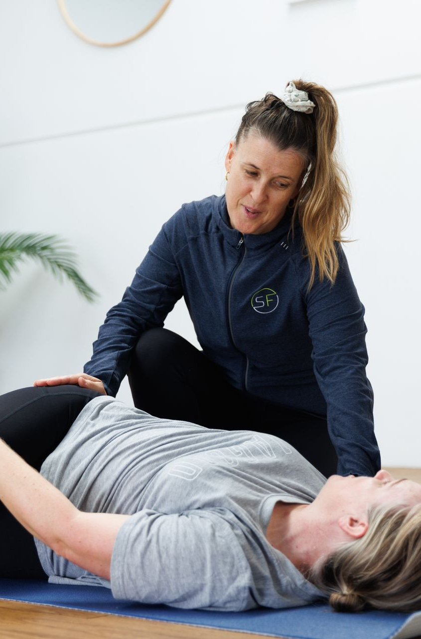 Woman assists another during a floor exercise in a studio. The woman kneels, hands on the other's abdomen.