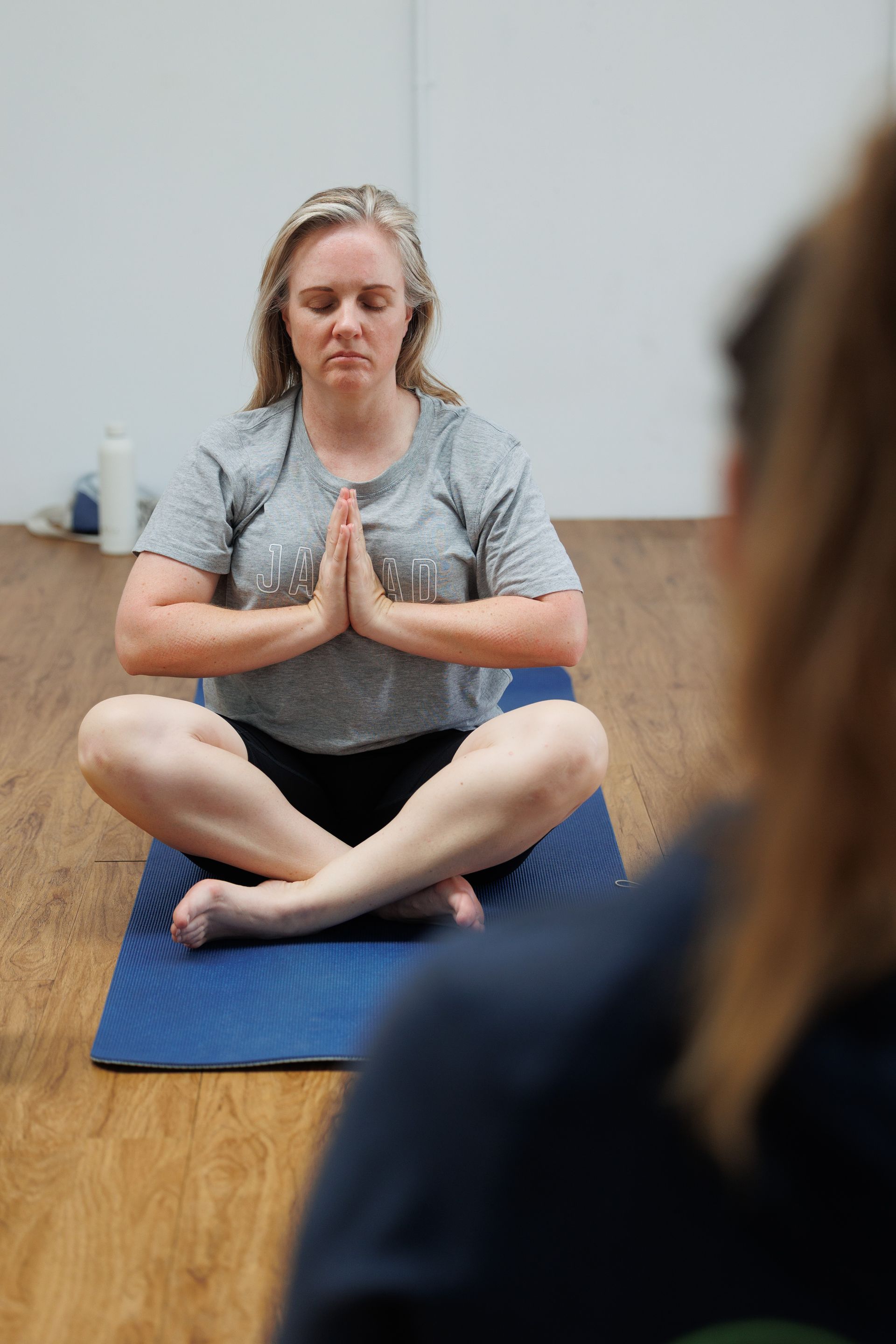 Woman meditating on a blue mat, hands in prayer pose, eyes closed, in a studio with a wooden floor.