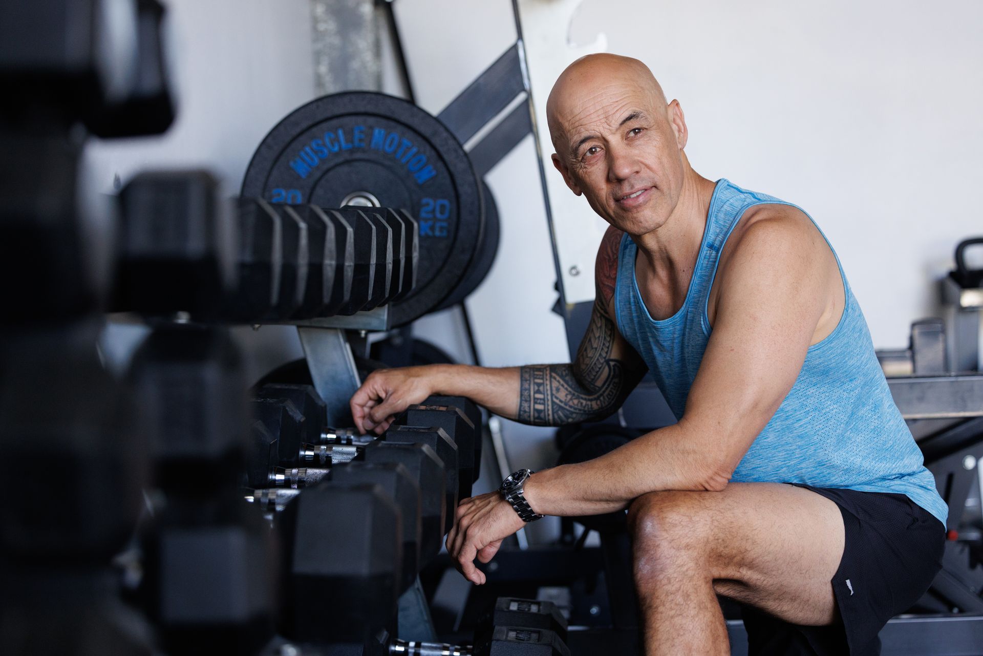Man in gym, resting arm on dumbbell rack, looking at the viewer. He wears blue tank top and black shorts.