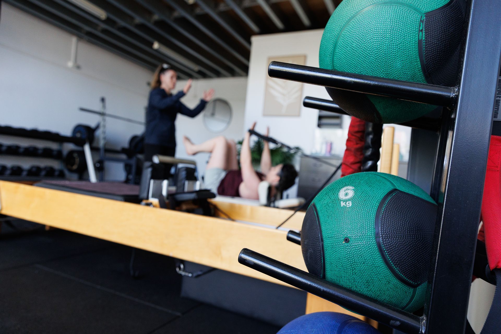 Rack with green medicine balls in foreground; person exercising with trainer in background at gym.