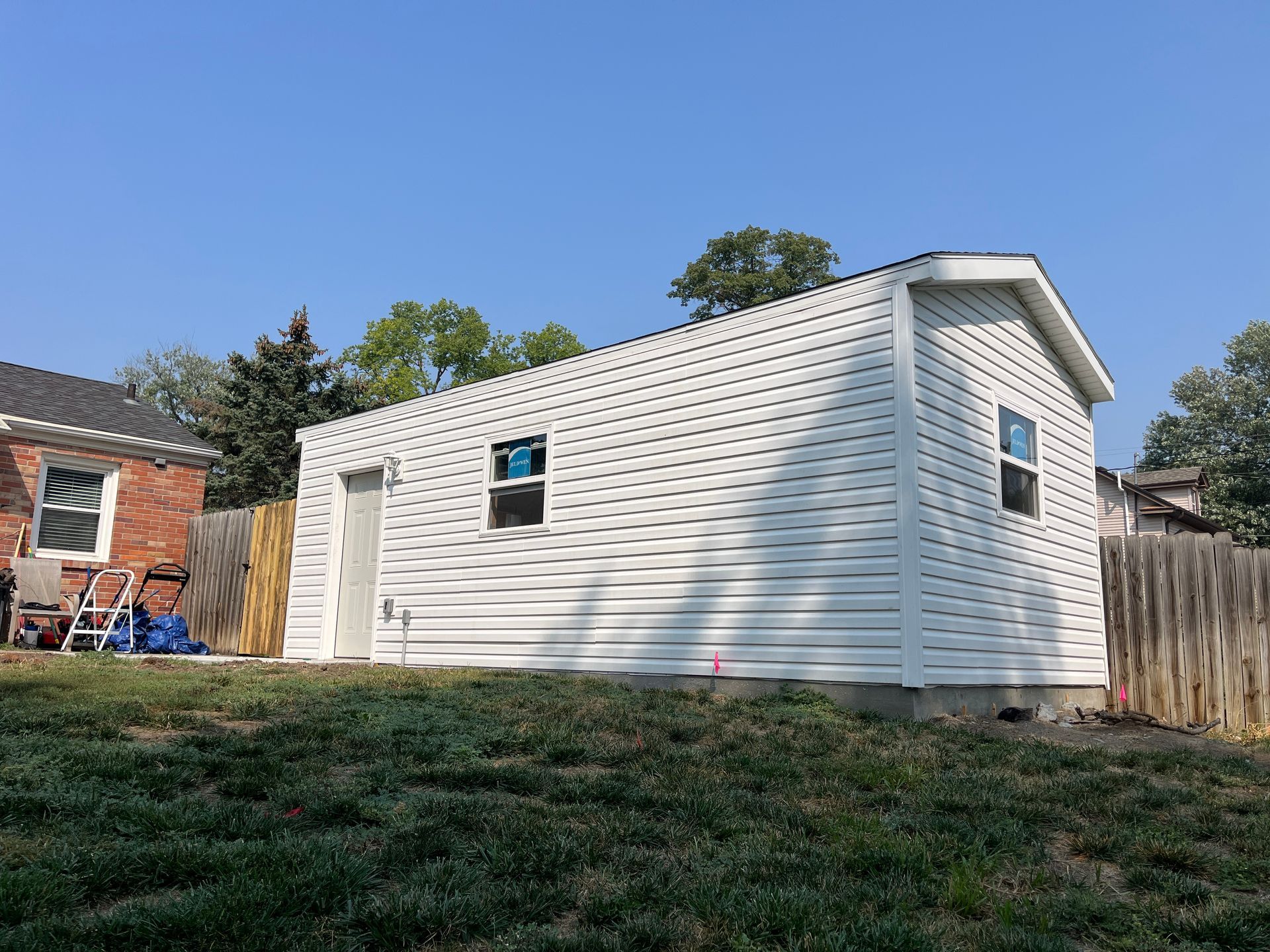 White vinyl-sided shed under construction, with two windows, door, and dark roof, on a grassy hill.