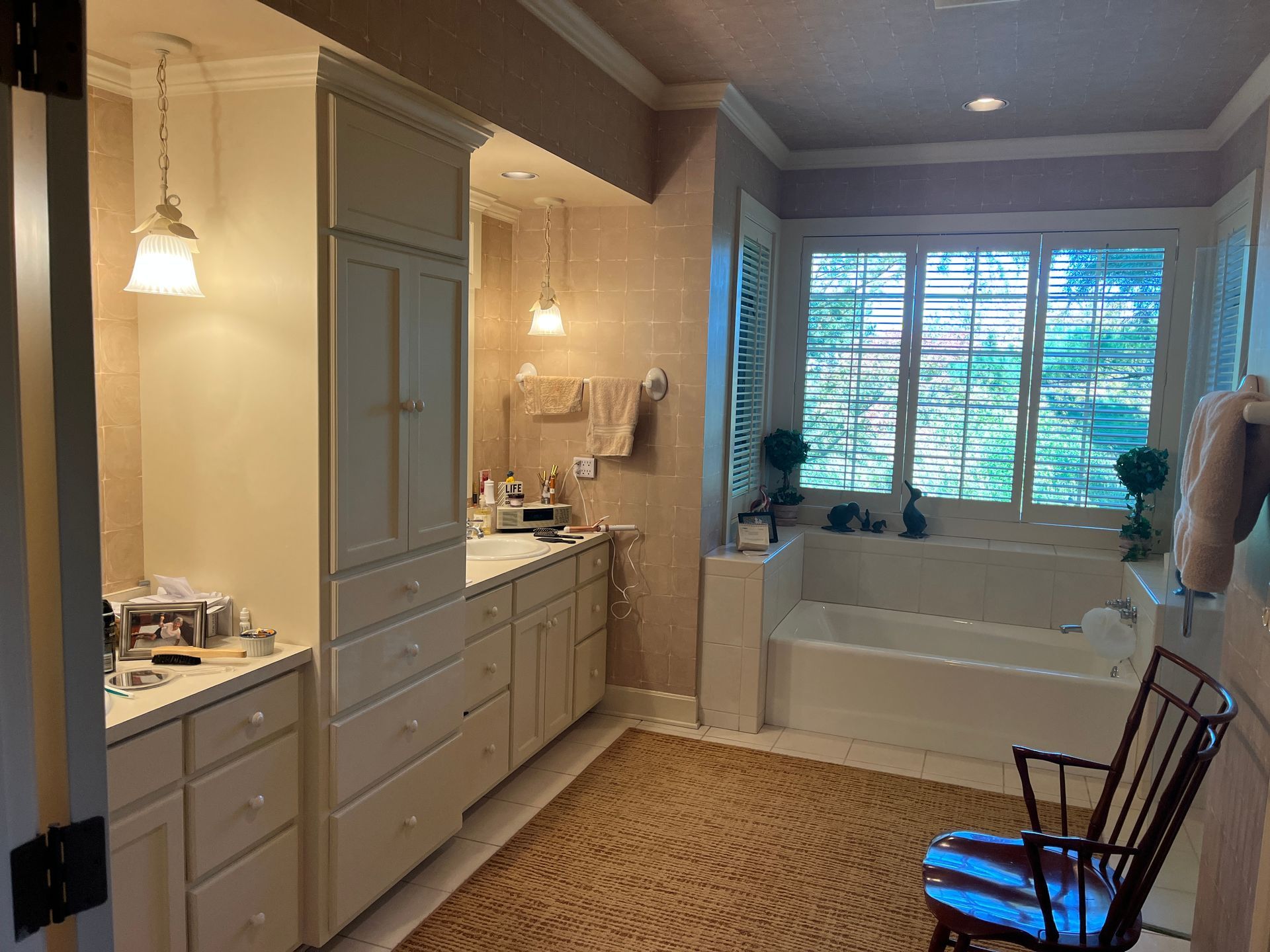 Bathroom with white cabinets, bathtub by window with shutters, beige rug.
