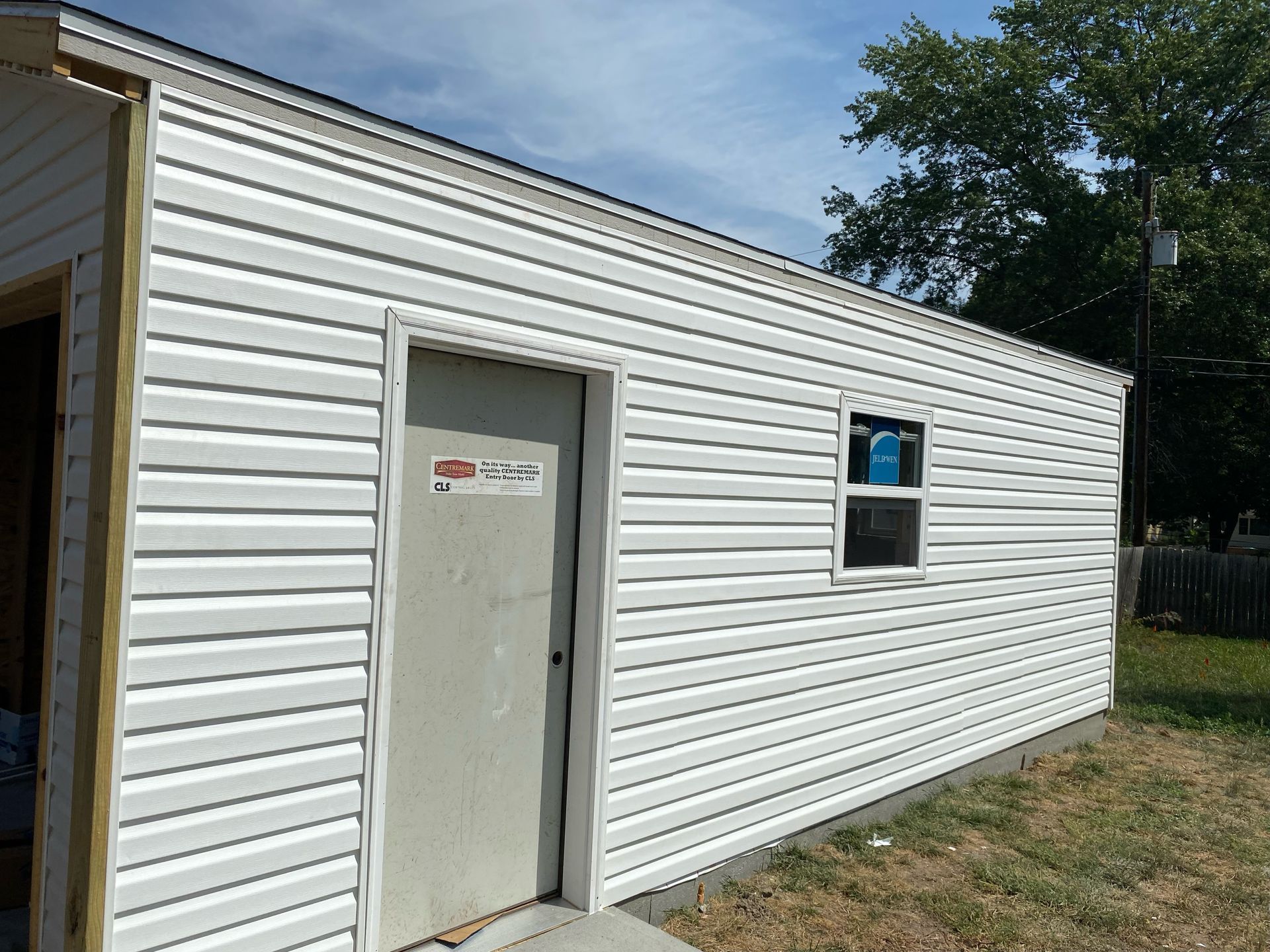 White-sided building with a door and window, set against a blue sky and green trees.