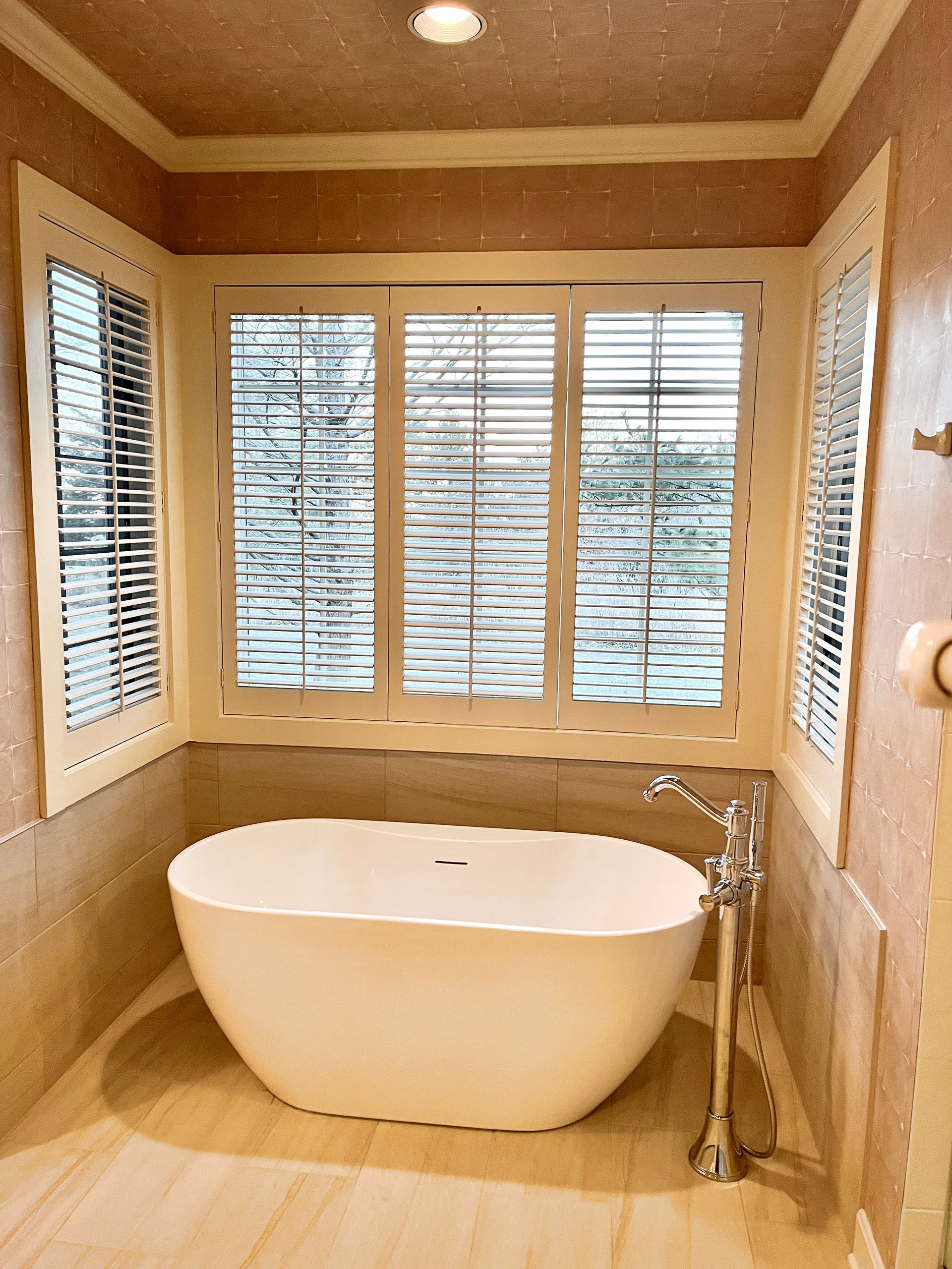 A white tub sits in a bathroom with large windows, beige walls, and blinds.