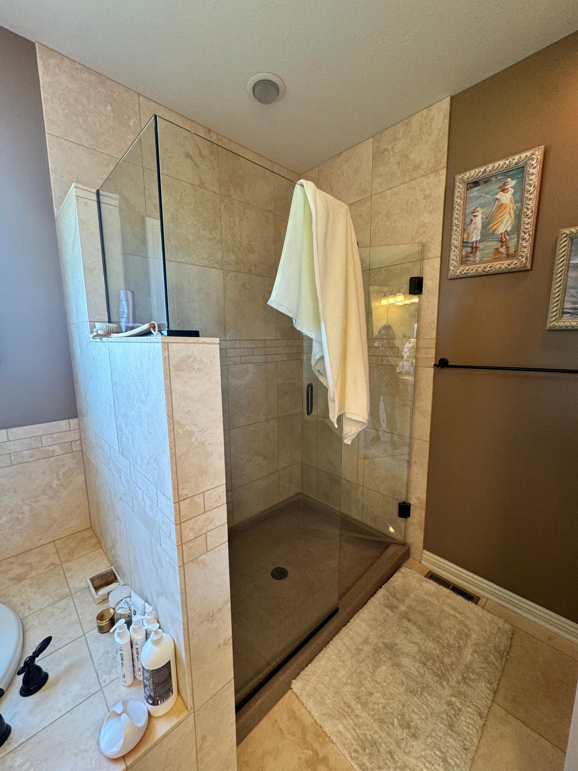 A beige tiled shower with glass door and a towel hanging inside. A rug sits outside the shower.