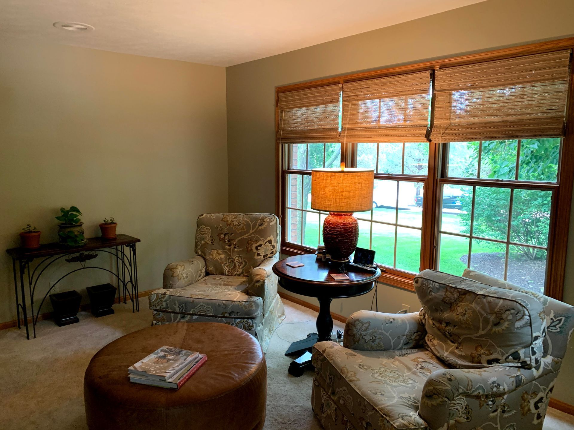 Cozy living room with two armchairs, a round ottoman, table, and a window with shades, near a lamp.