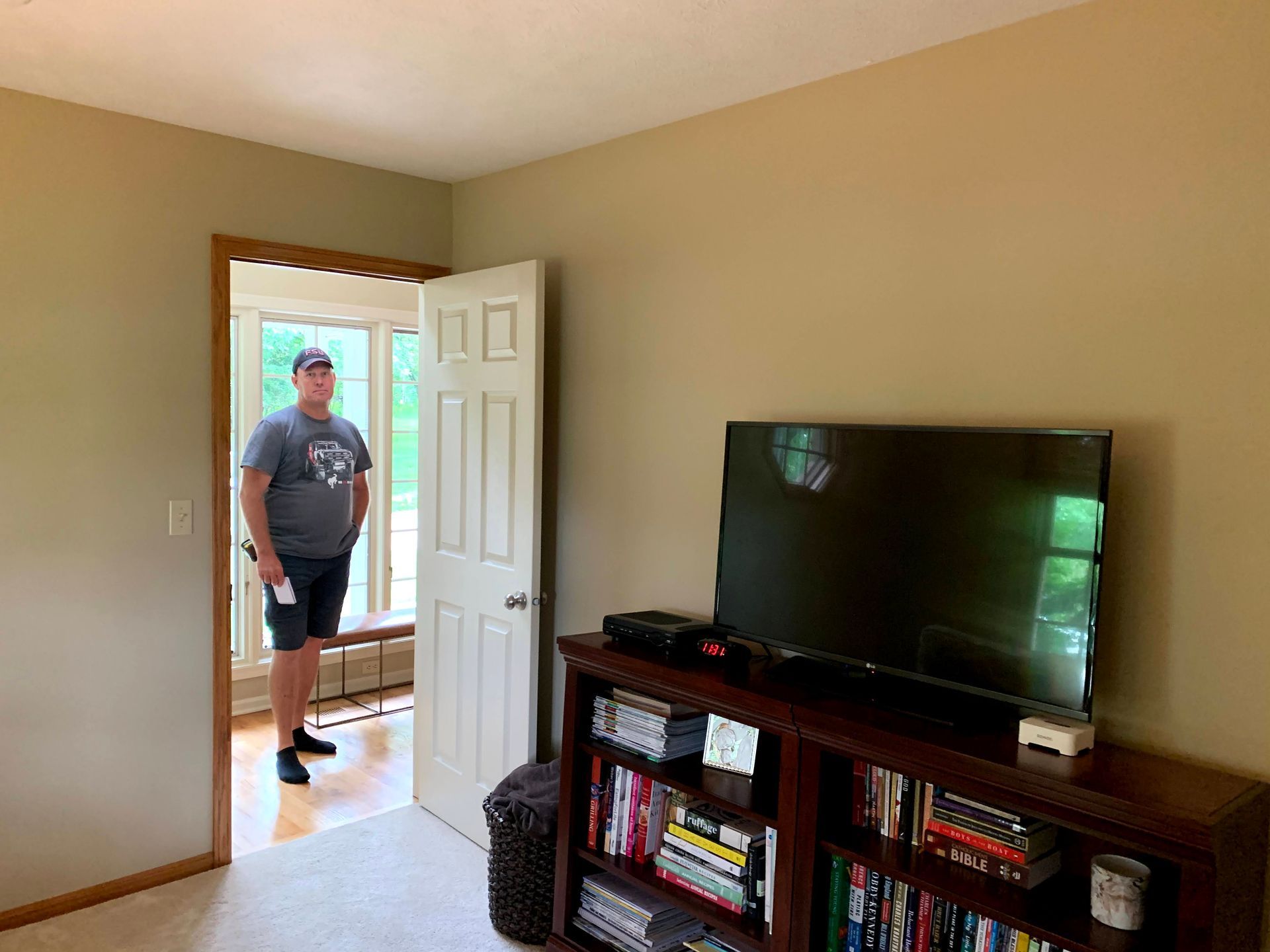 Man in a room, standing in a doorway with a TV on a bookshelf. Beige walls, carpet, and natural light.