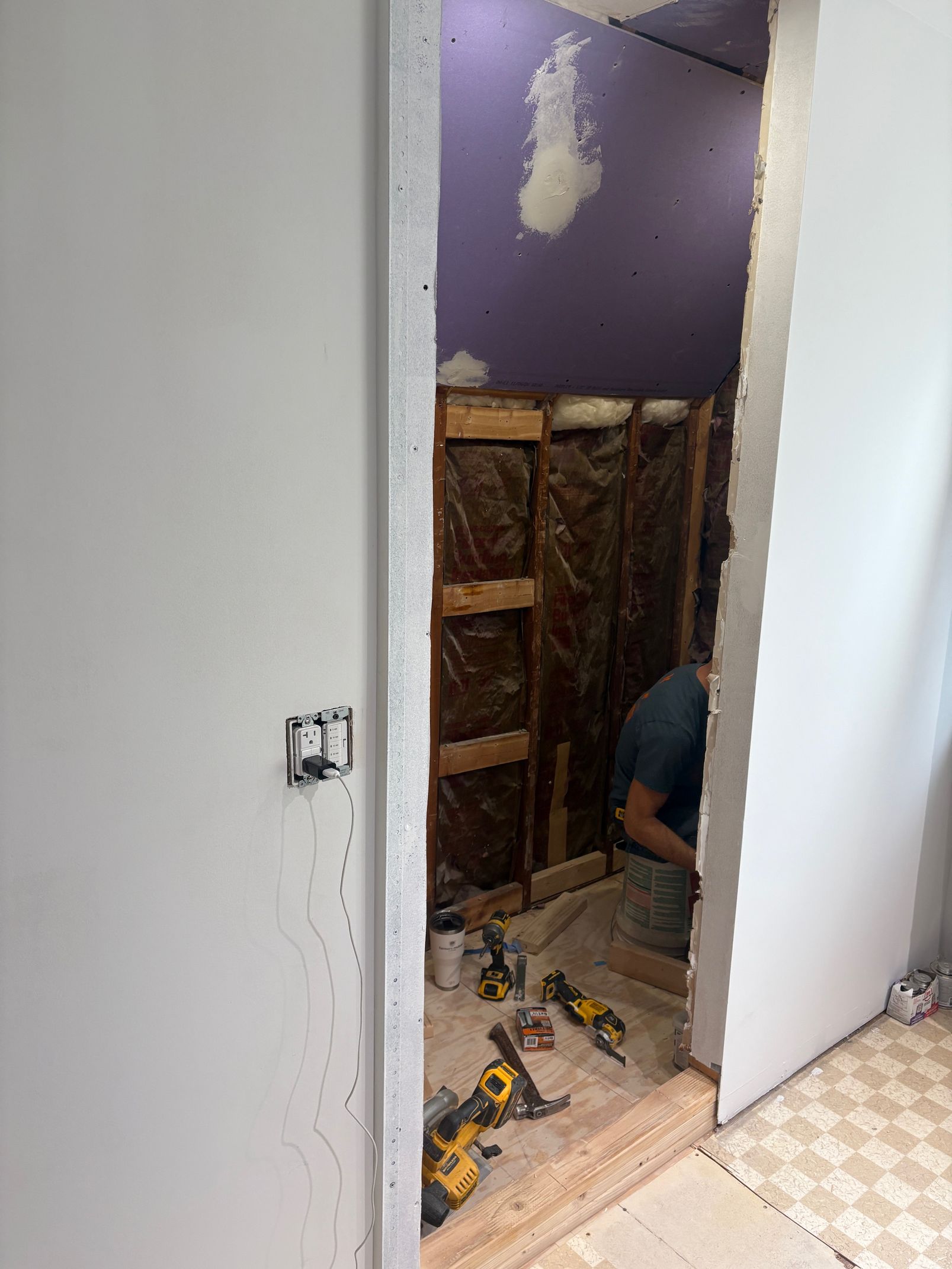 Open doorway into a bathroom under renovation; a worker is visible, tools on the floor, exposed framing, and purple wall.