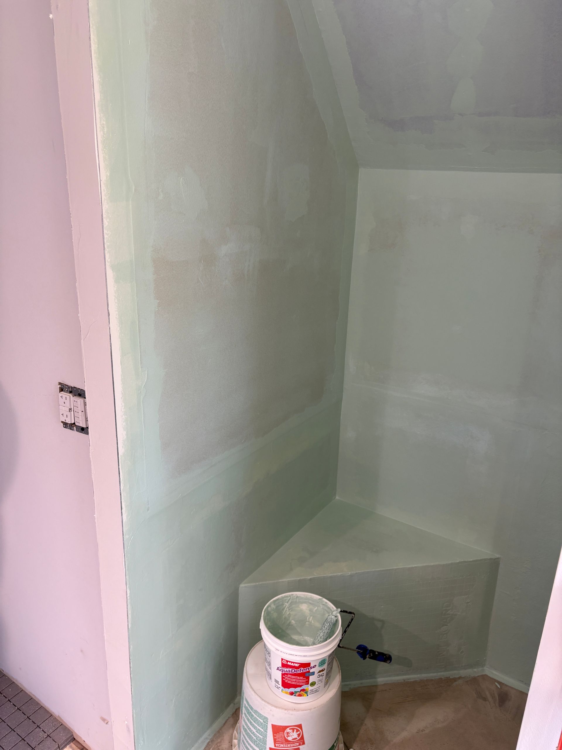 Green-painted bathroom corner with bench. A bucket of joint compound sits below. Walls, in the process of being finished.