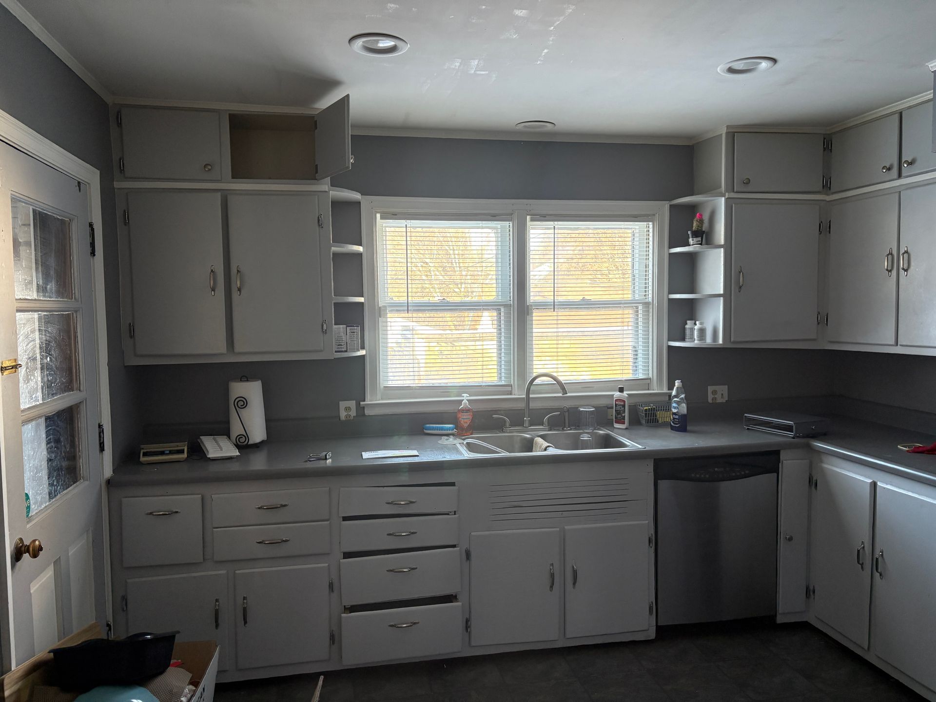 A small kitchen with white cabinets, gray counters, and a window above the sink.