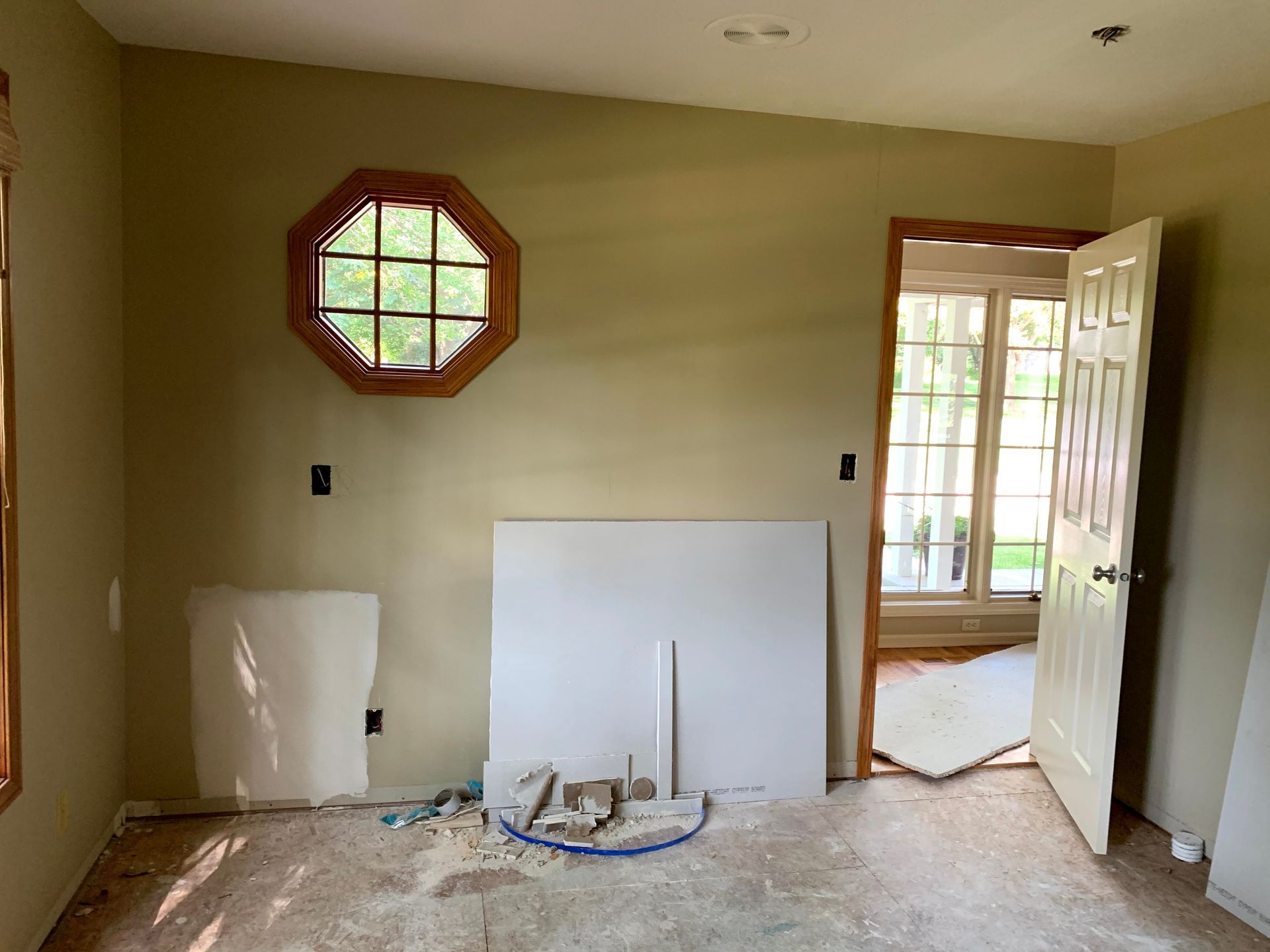 Interior room under renovation: tan walls, octagonal window, door open, drywall leaning against wall, debris on floor.