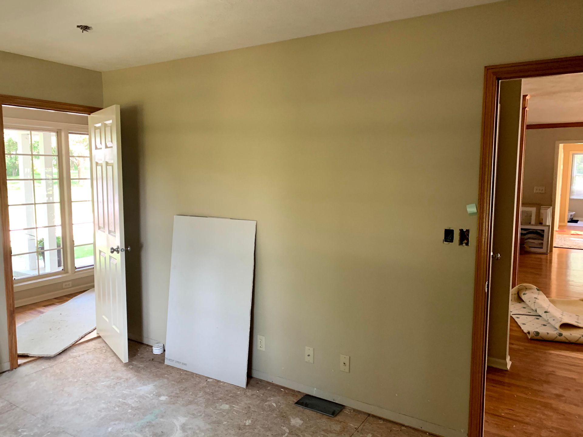 Room with beige walls, open door to a sunny outdoor view, and another door leading to a room with hardwood floors.