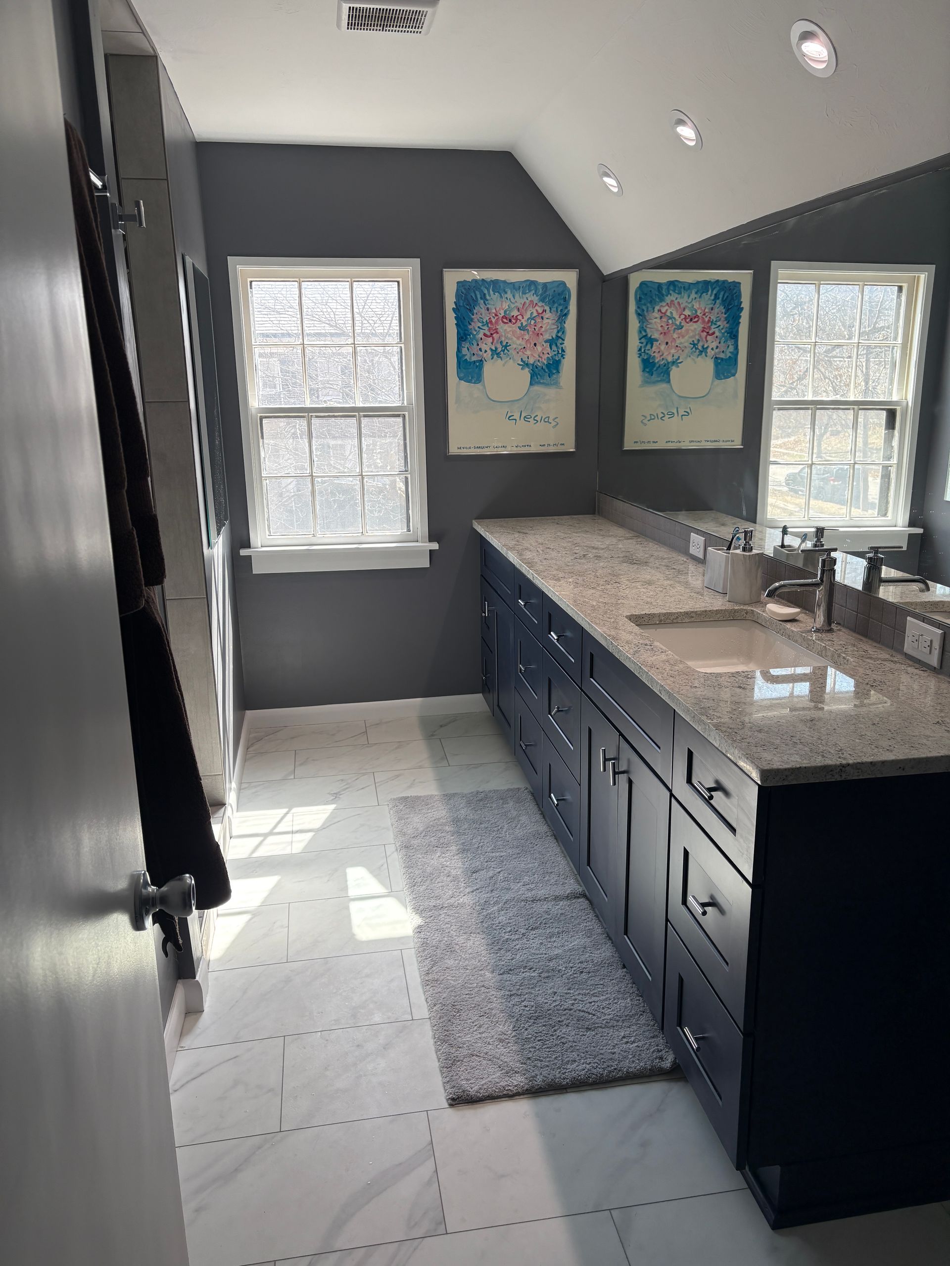 Dark blue bathroom with two windows, granite countertop, two framed prints and a rug.