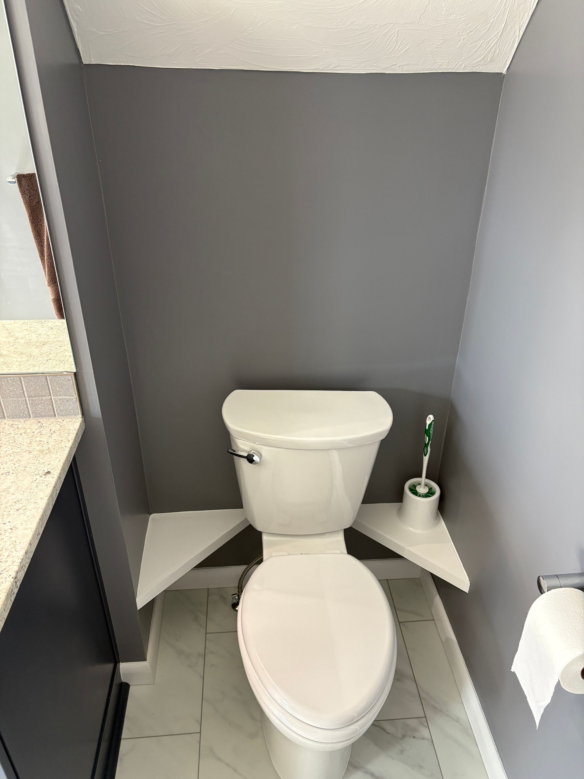 Toilet in a small bathroom, with grey walls, white shelves, and a white toilet paper roll.