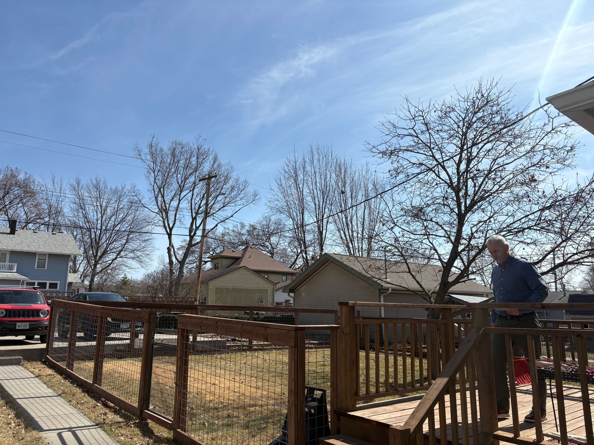 Man on a wooden deck overlooking a yard with bare trees and houses under a blue sky.