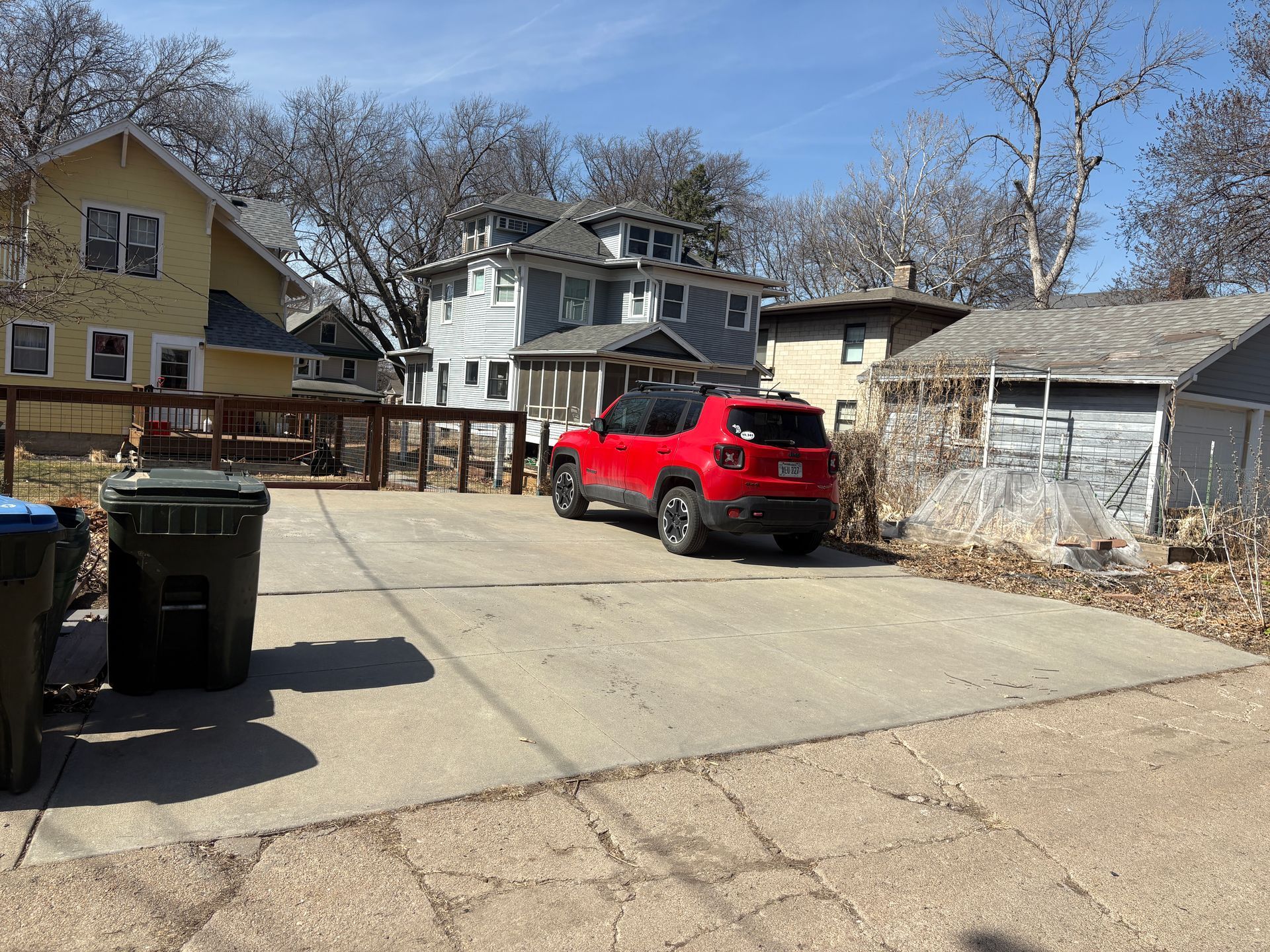 Red Jeep parked on concrete driveway in neighborhood, next to trash cans and wooden buildings.