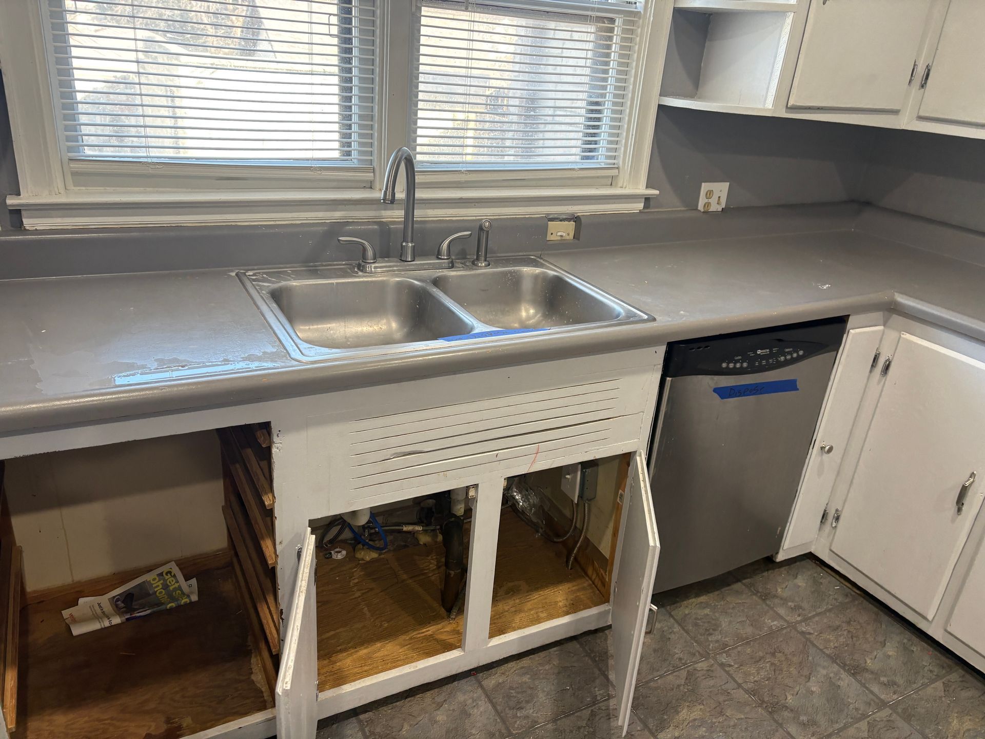Kitchen with open cabinet doors under a double sink. Dishwasher to the right. Gray countertops.