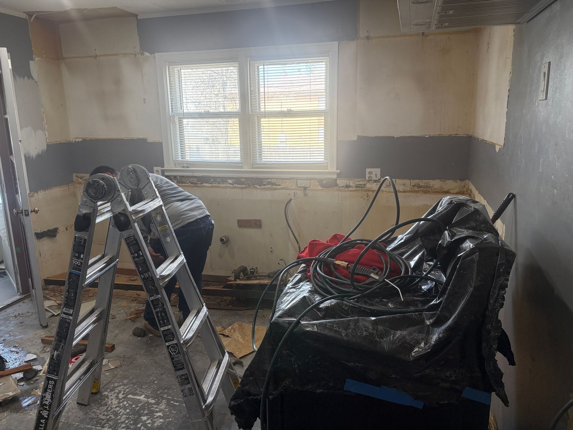 Kitchen under renovation; person working near a ladder. Walls are exposed, window in the center.