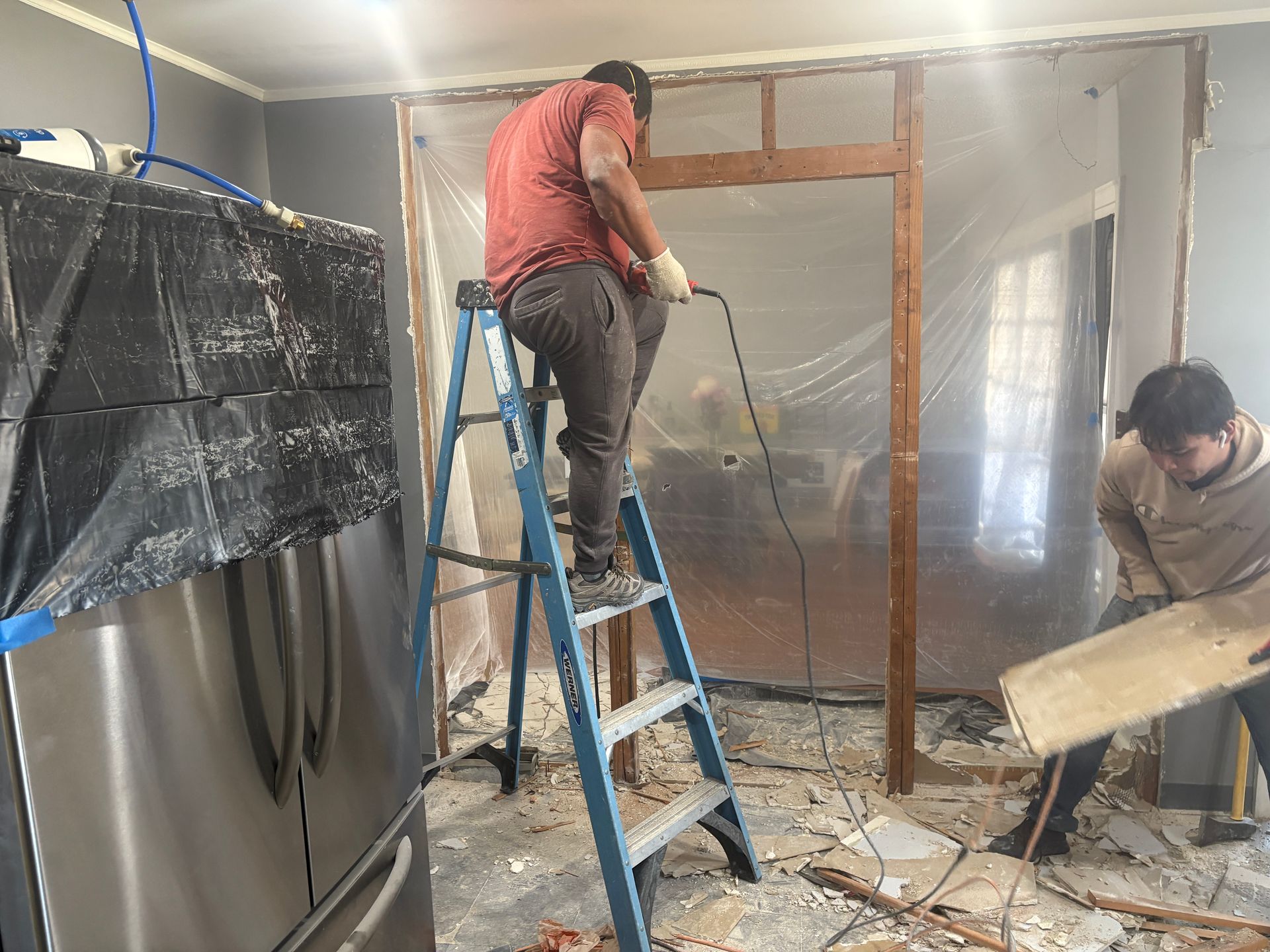 Two workers demolishing a doorway; one on a ladder using a tool, the other holding a board. Construction in progress.