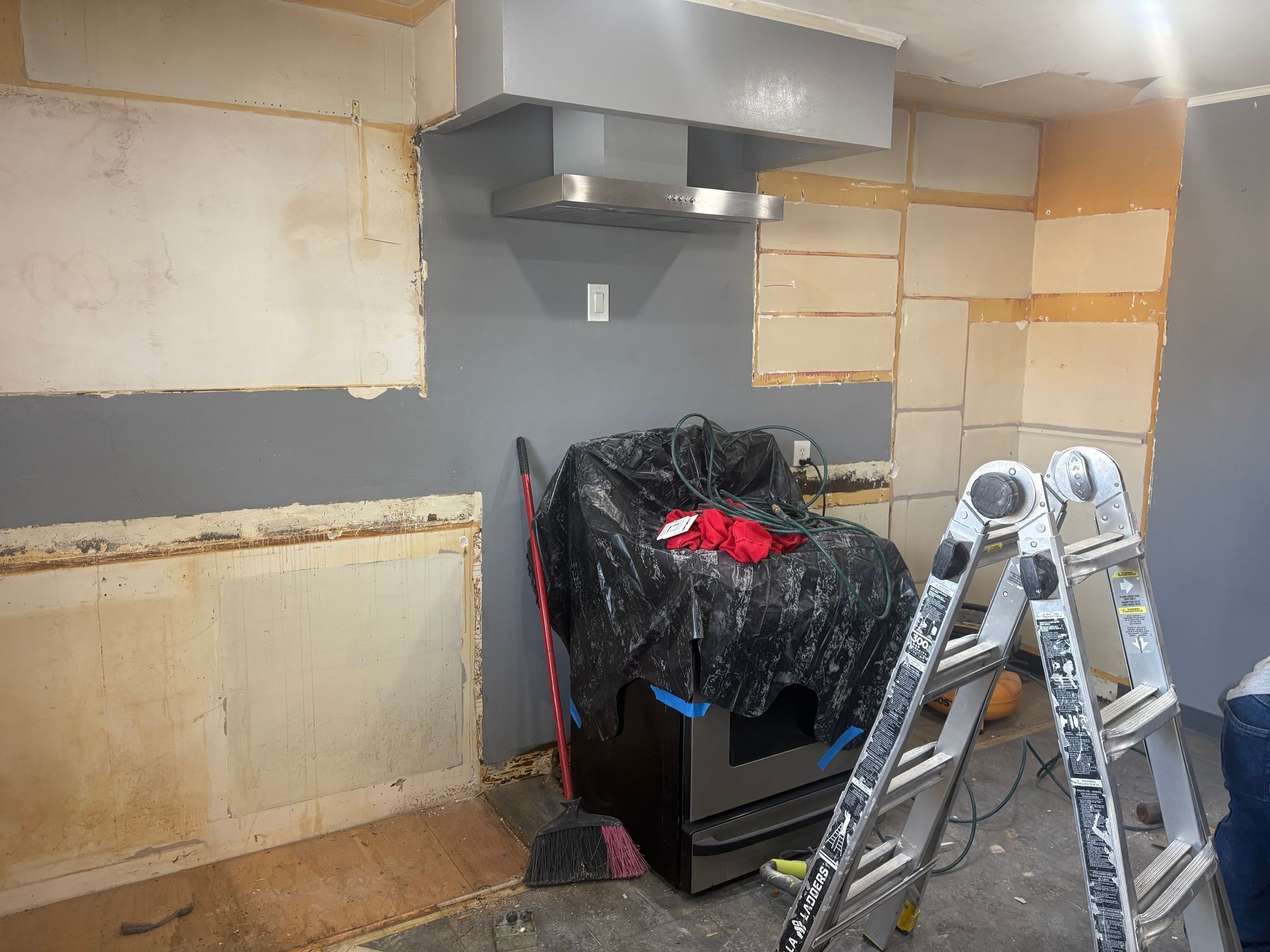 Kitchen under renovation with range, hood, and ladder. Gray and exposed wall sections.