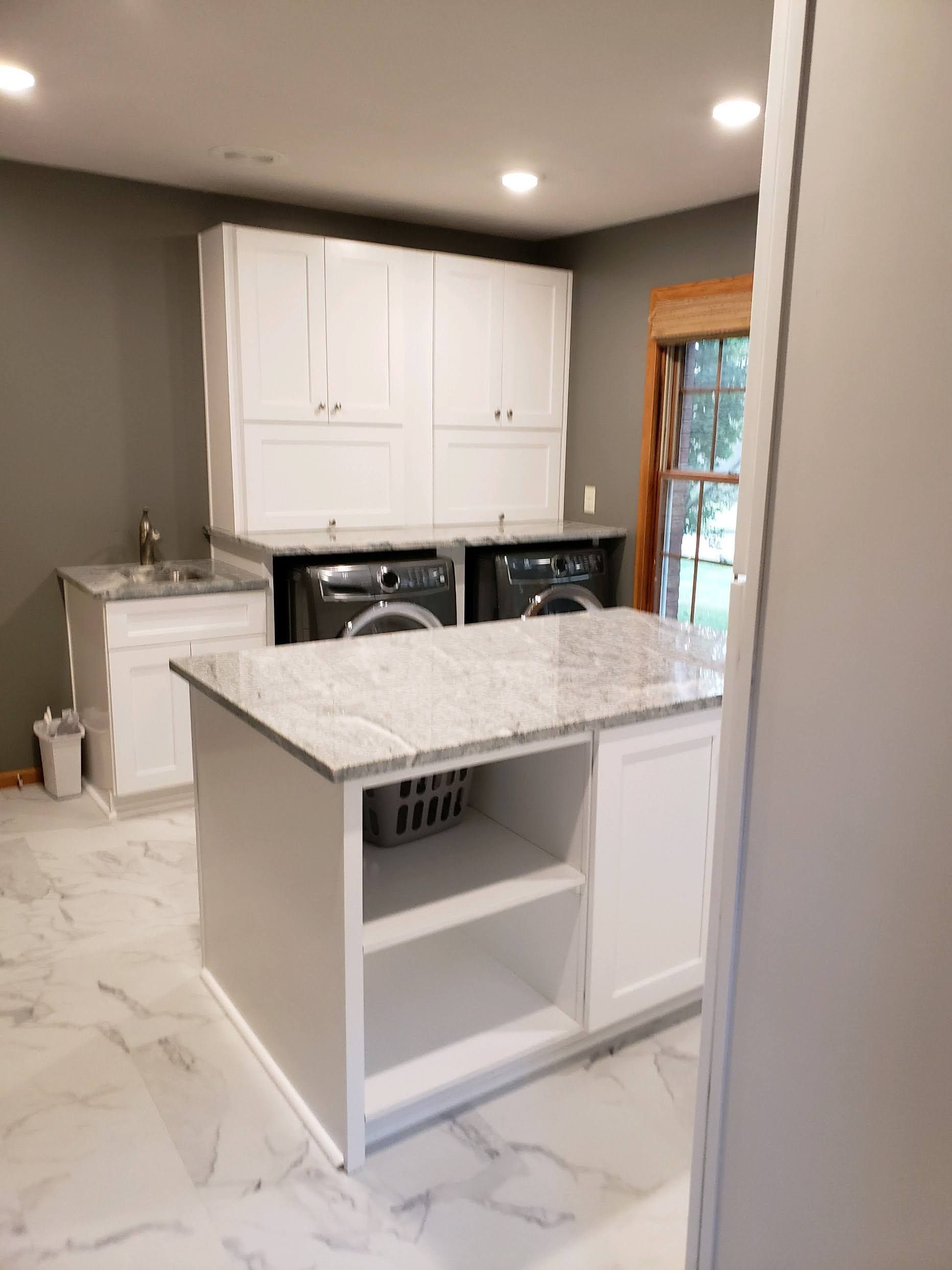 Modern laundry room with white cabinets, island, and washer/dryer. Granite countertops and marble-look floor.