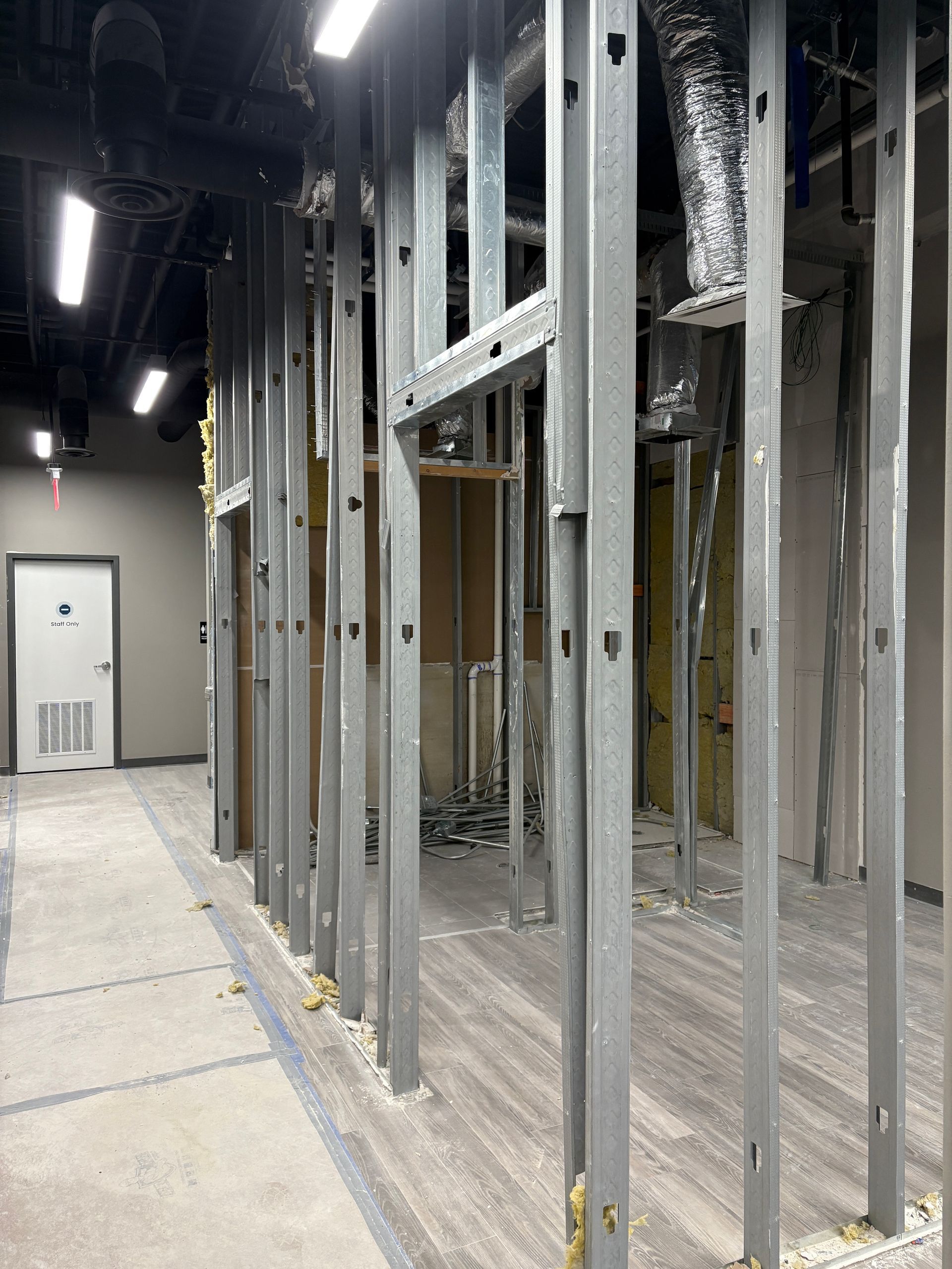 Construction site: metal studs framing a wall with doorway, hallway, HVAC, and a door in the distance.