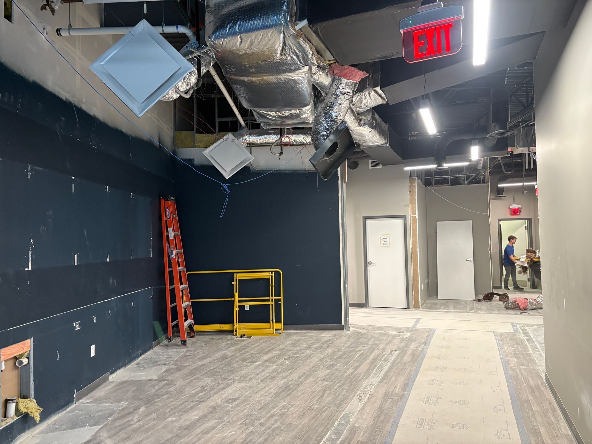 Interior construction site: dark blue wall, exposed ductwork, ladder, yellow lift, workers.