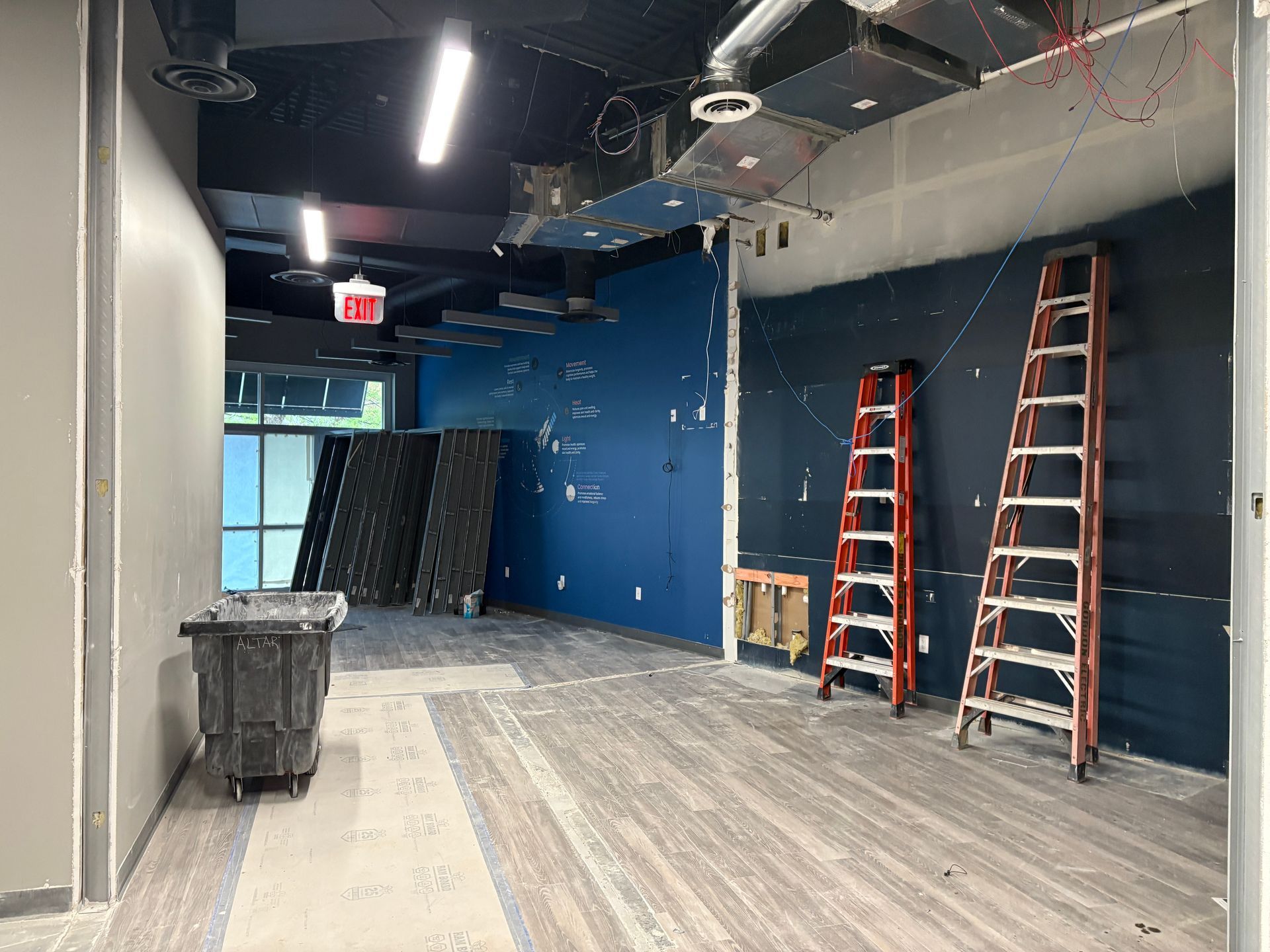 Interior construction site. Dark blue wall with two ladders, trash bin, and exposed ceiling.