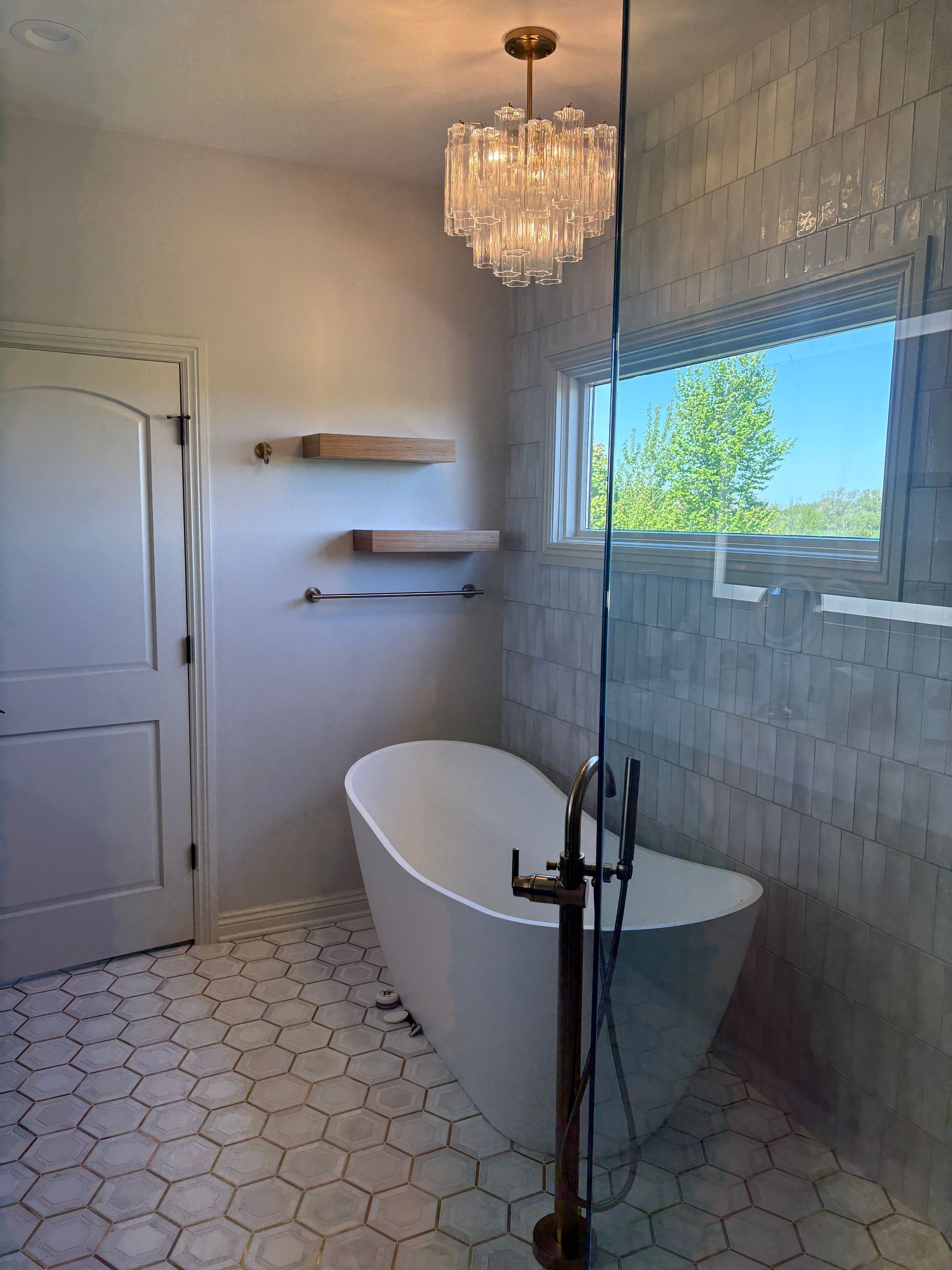 Bathroom with a white bathtub, chandelier, shelves, window, and hexagon tile floor.