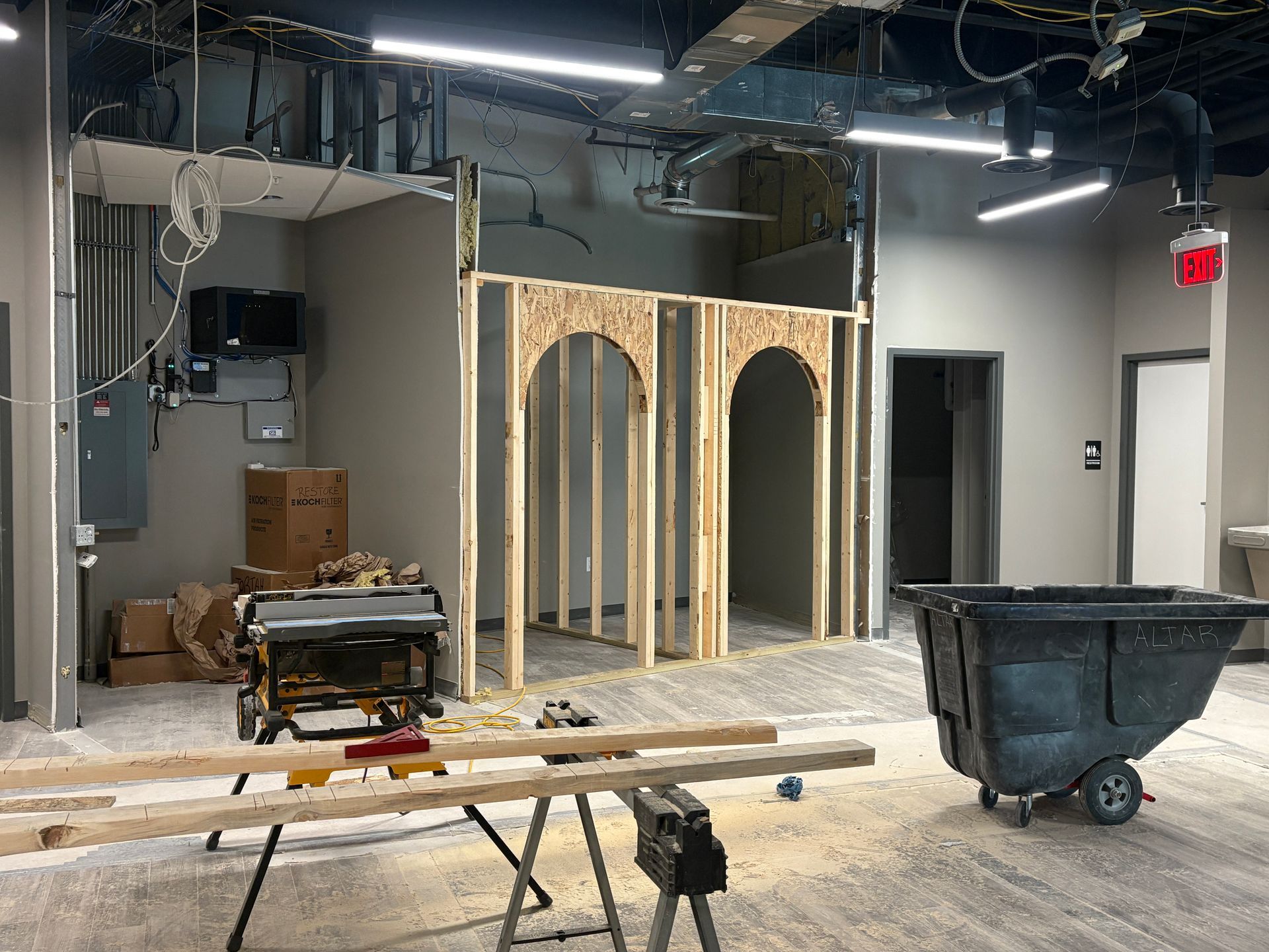 Construction site: Interior view with wood framing, tools, and debris. A black trash bin is in the foreground.