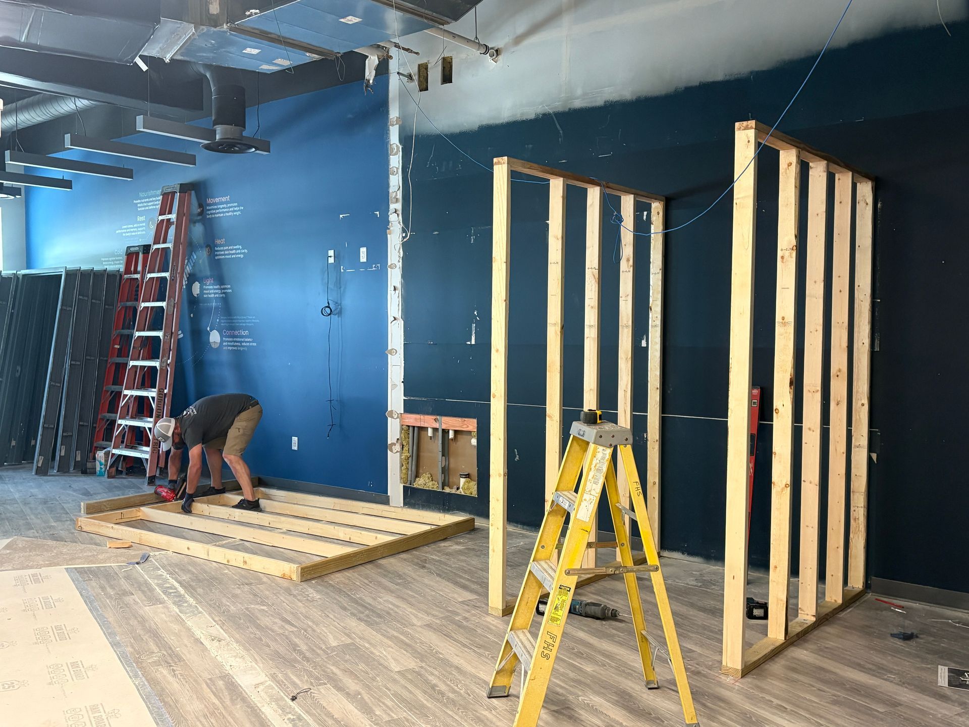 Construction site: man building a wall with wood. Blue walls and a ladder are visible.