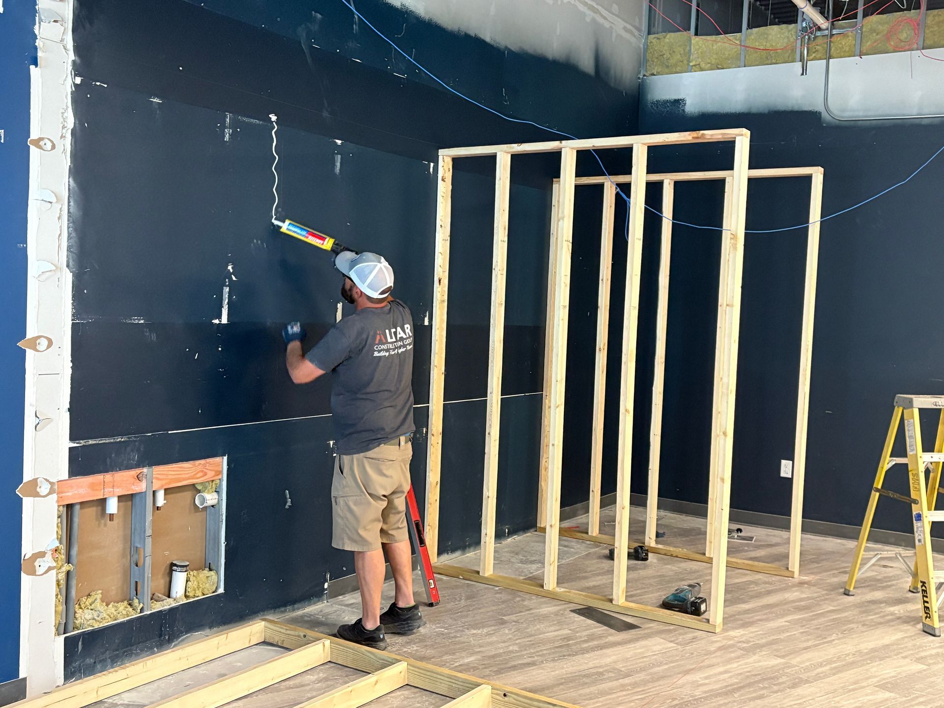 A person uses caulk on a dark blue wall, with wood framing nearby, construction site.