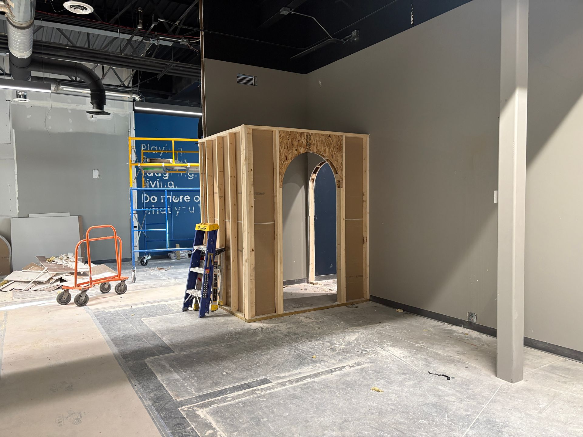 Construction in a room: wood frame structure, gray walls, concrete floor, scaffolding visible.