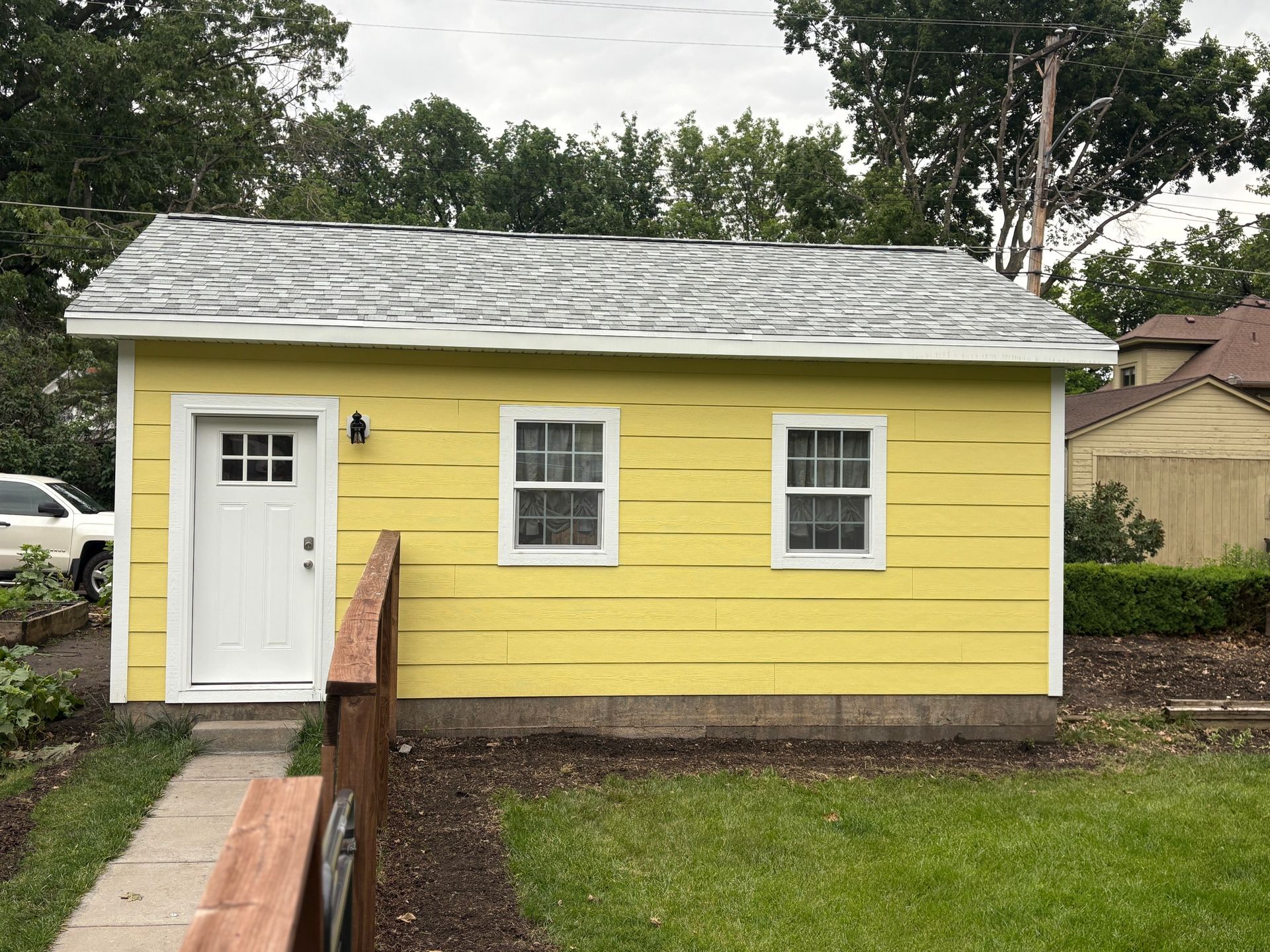 Yellow shed with white door and windows, gray roof, and wooden steps in a grassy yard.
