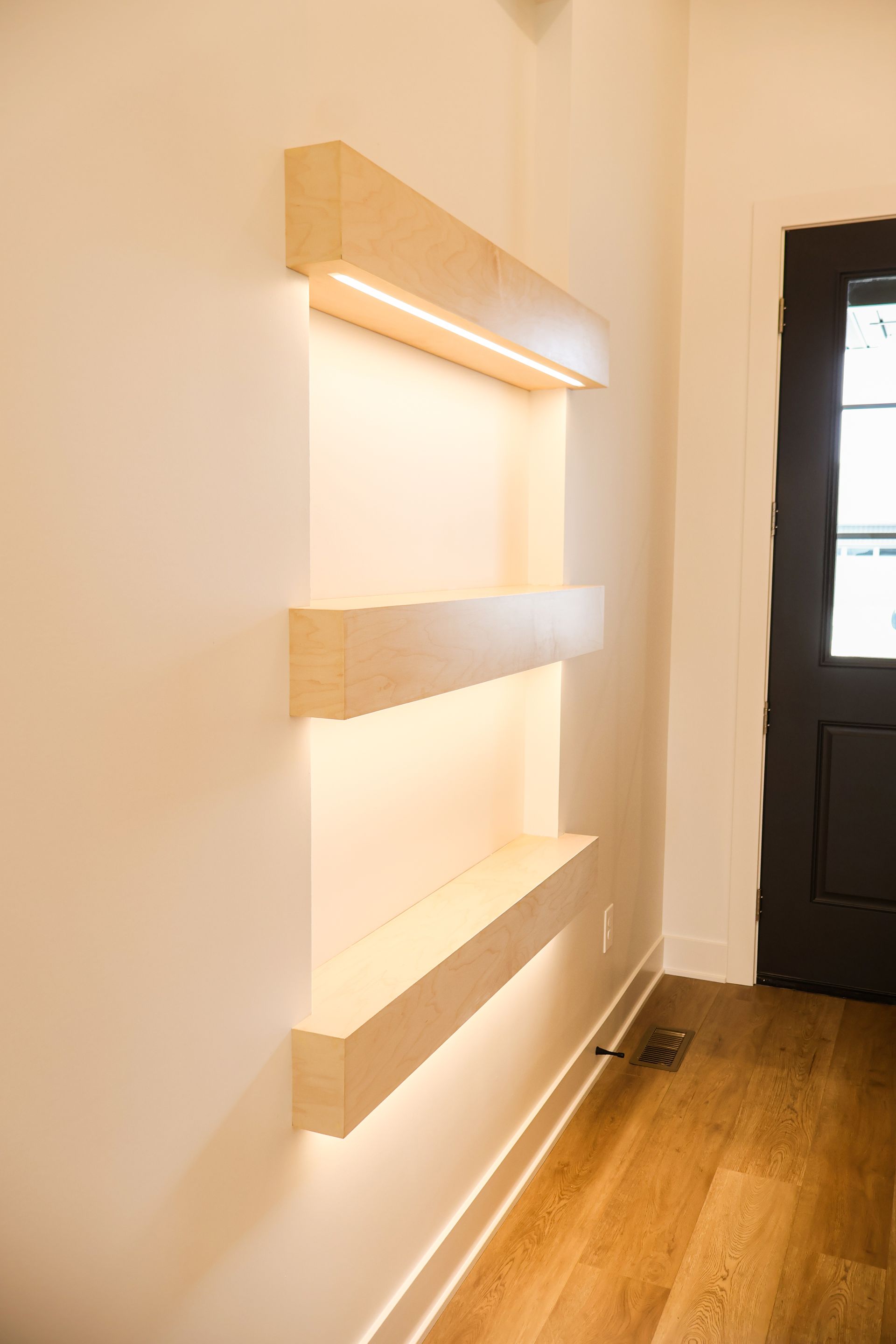 Three modern, rectangular recessed shelves with built-in lights on a white wall, next to a black door.