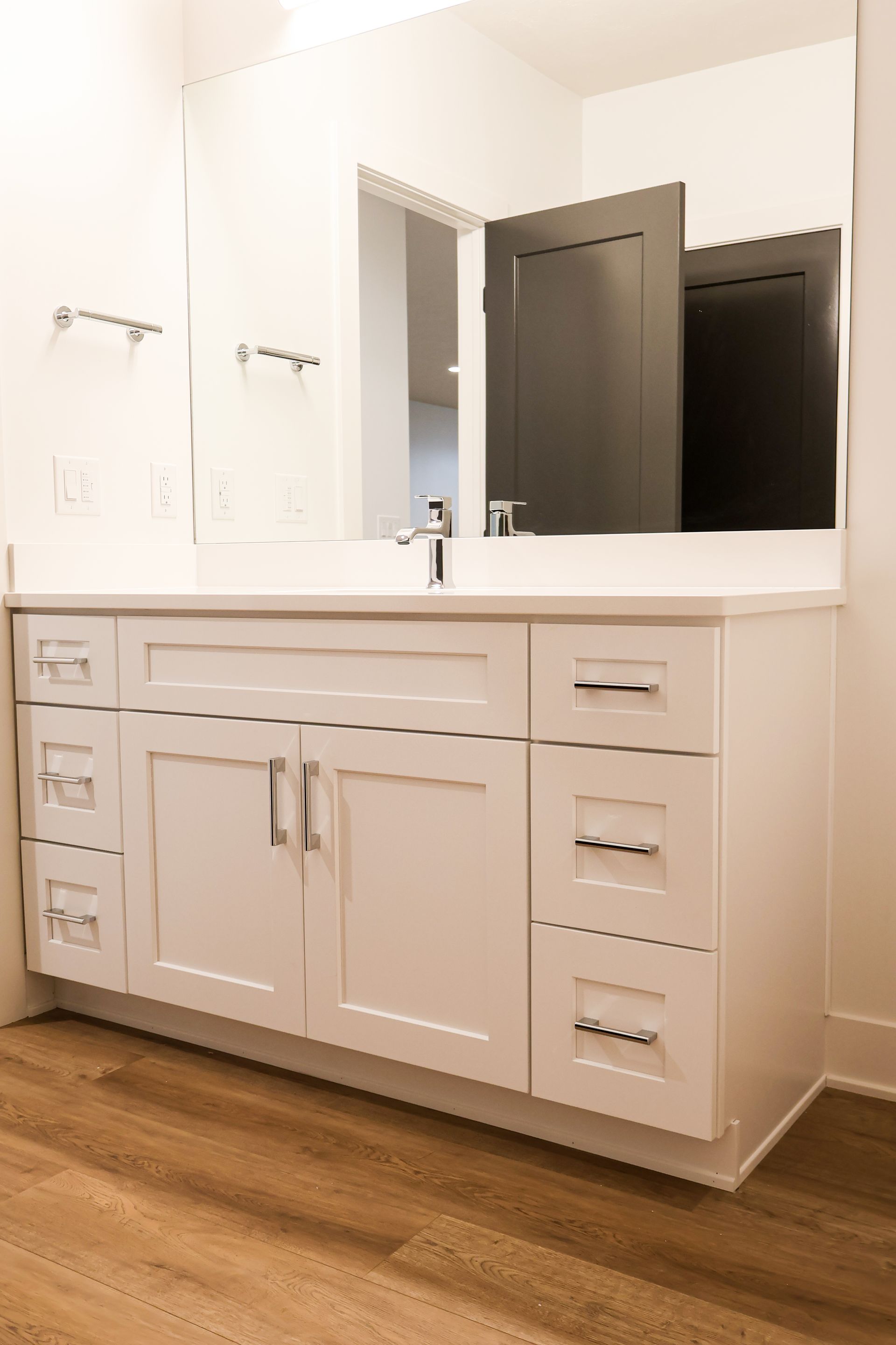White bathroom vanity with drawers and cabinets, chrome hardware, large mirror, and open door.