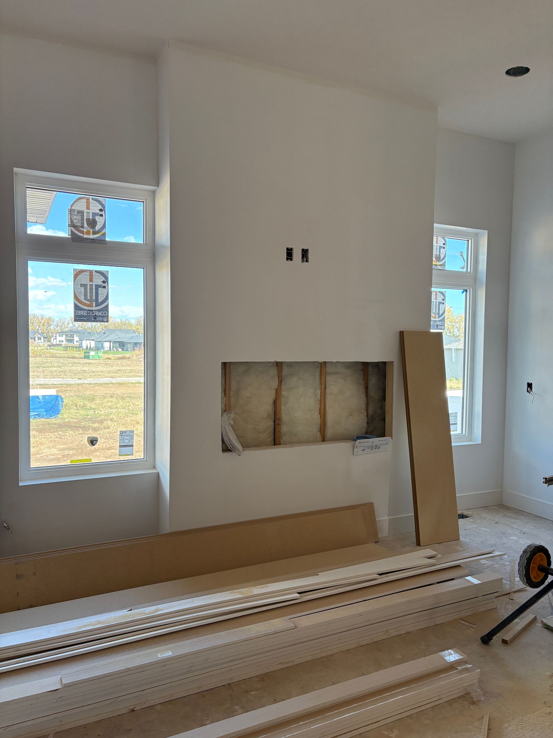 Interior of a room under construction with a fireplace and windows; white walls and trim, boards on the floor.