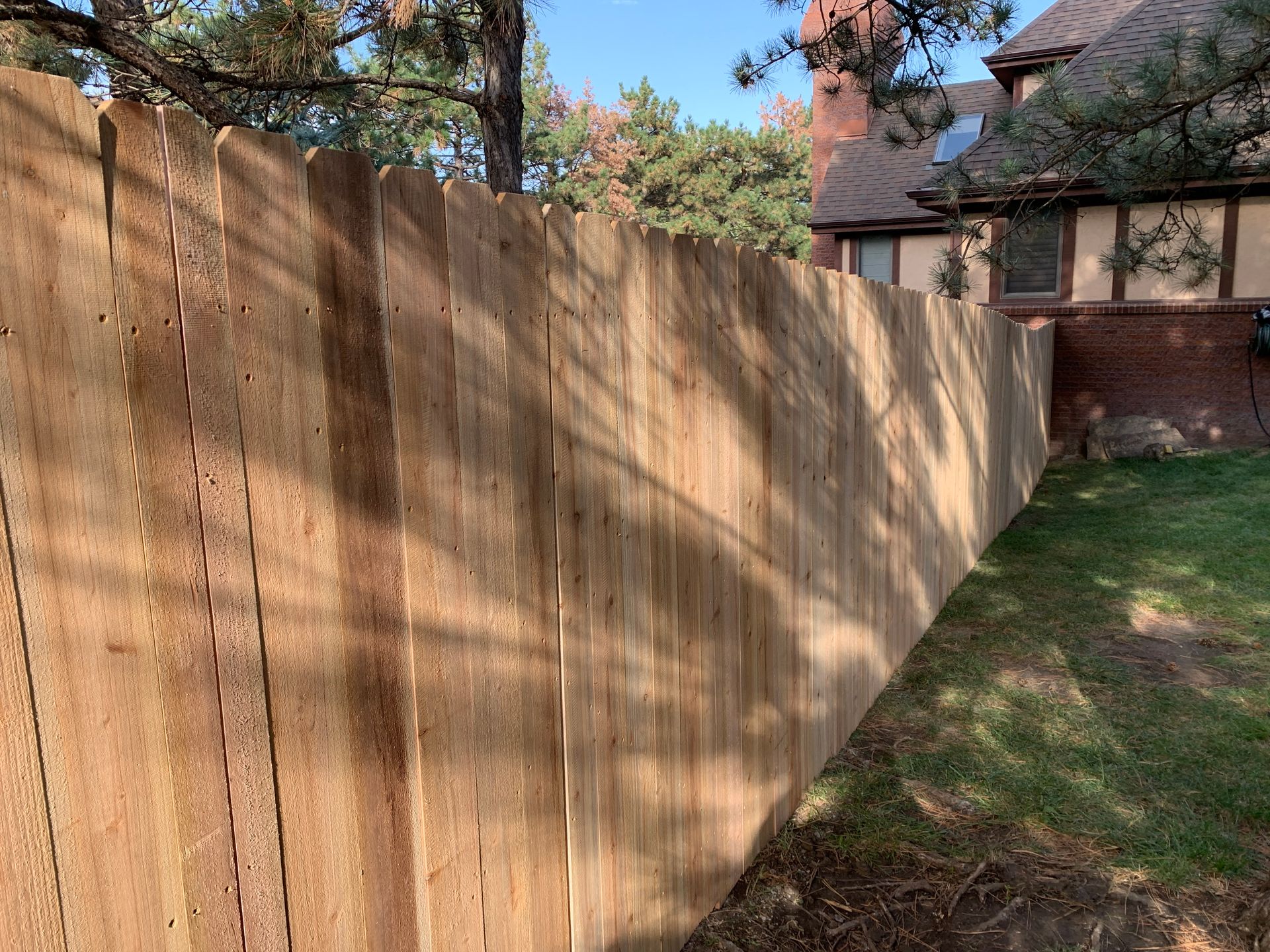 Wooden fence along a grassy yard, sunlight creating shadows. A house with brick and brown siding is visible in the background.