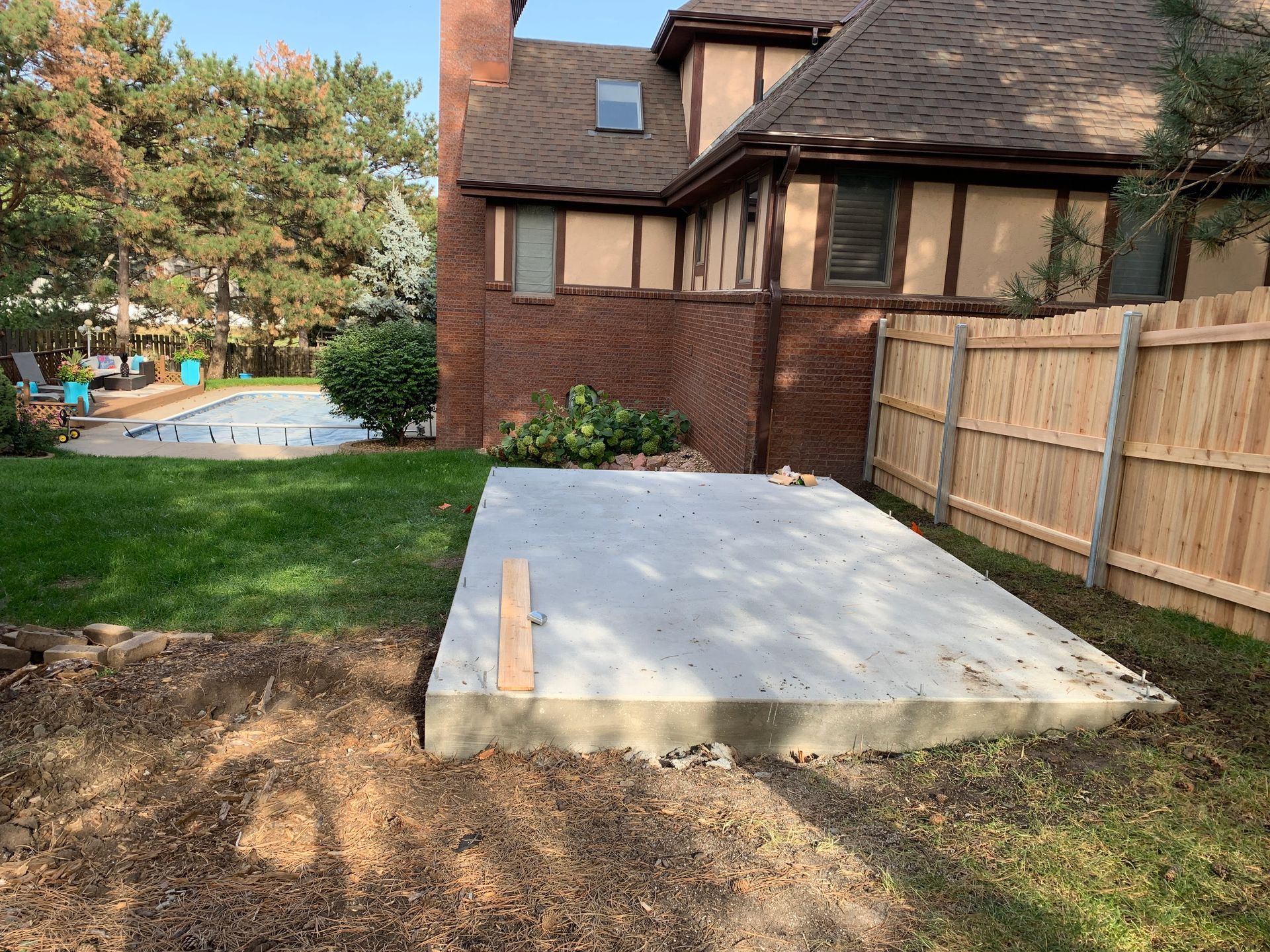 A newly poured concrete pad in a backyard, beside a wooden fence and a brick house. Green grass and trees surround it.