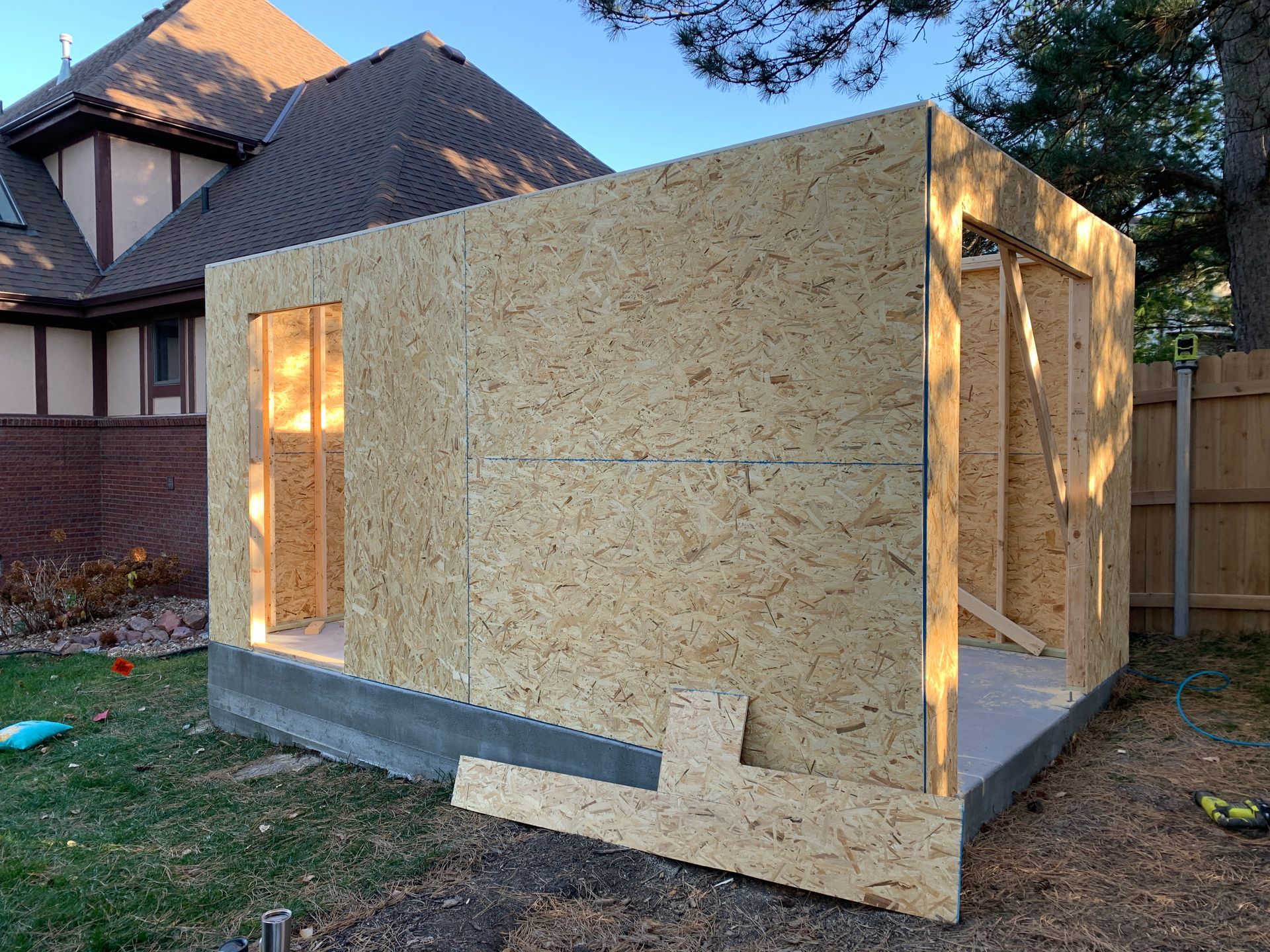 A partially built shed in a backyard. The structure is made of OSB panels on a concrete foundation, with openings for a door and window.