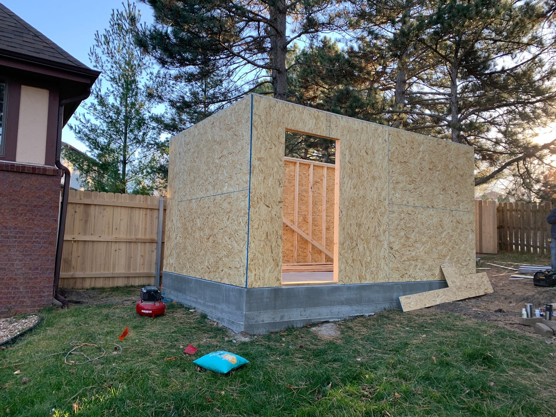 A partially built shed made of plywood, with a doorway and a concrete foundation, in a backyard.