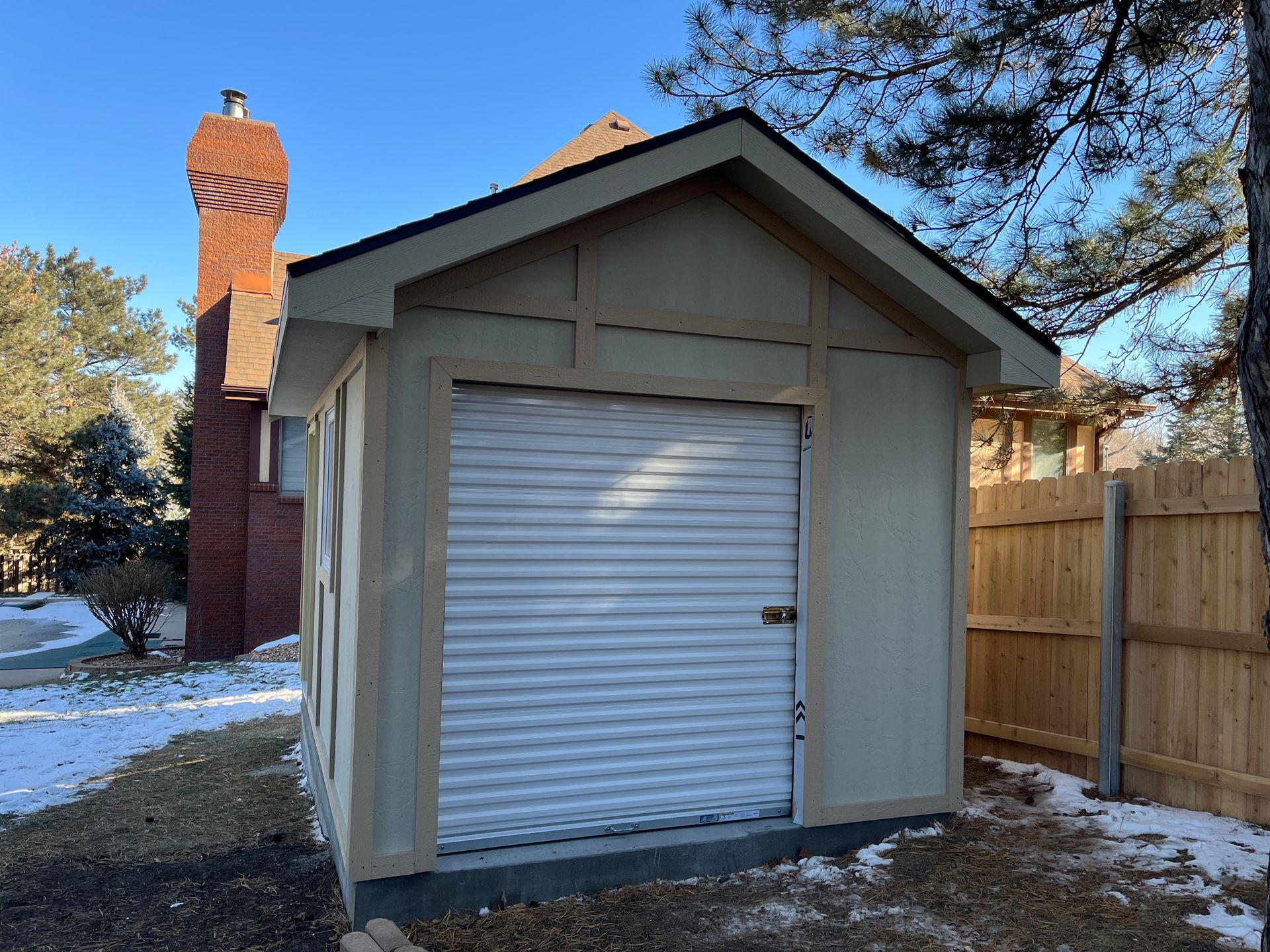Small beige shed with a roll-up door, in a snowy yard, beside a wooden fence and a brick chimney.